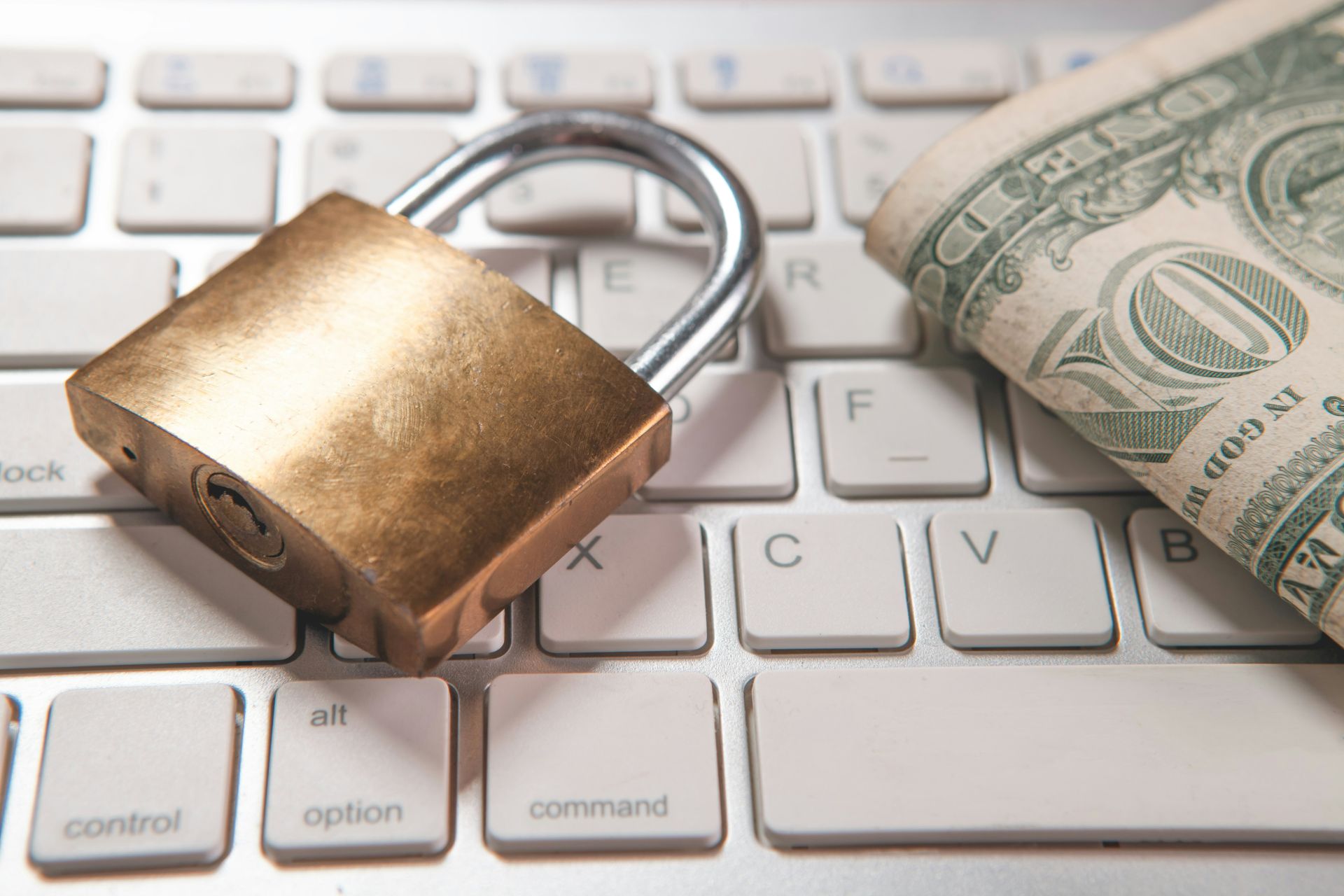 a gold padlock rests on a white keyboard next to a rolled U.S. dollar bill, symbolizing online security and financial protection.