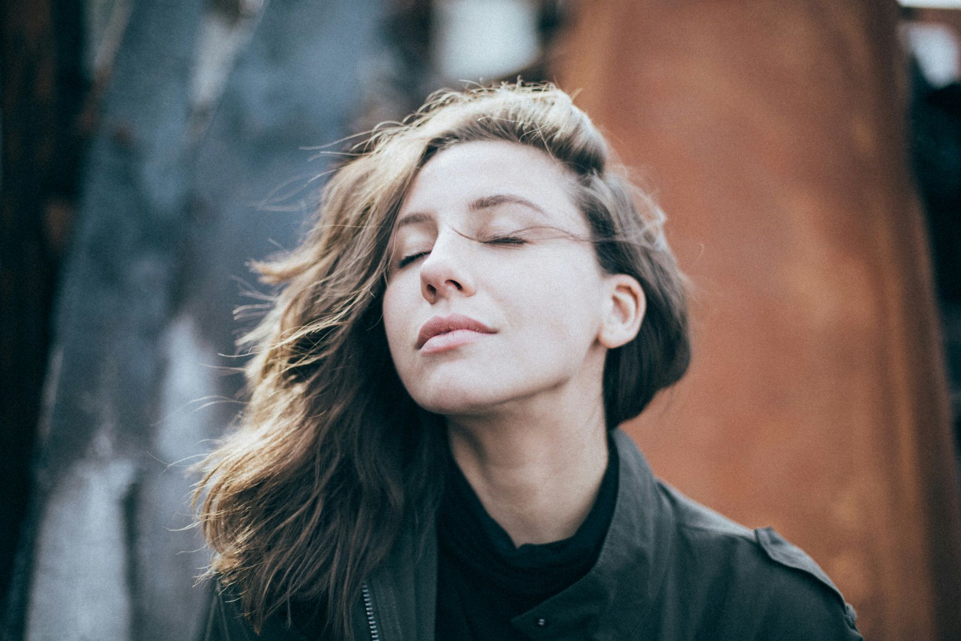 a woman with closed eyes and gently flowing hair stands outdoors, her face expressing calm and contentment.