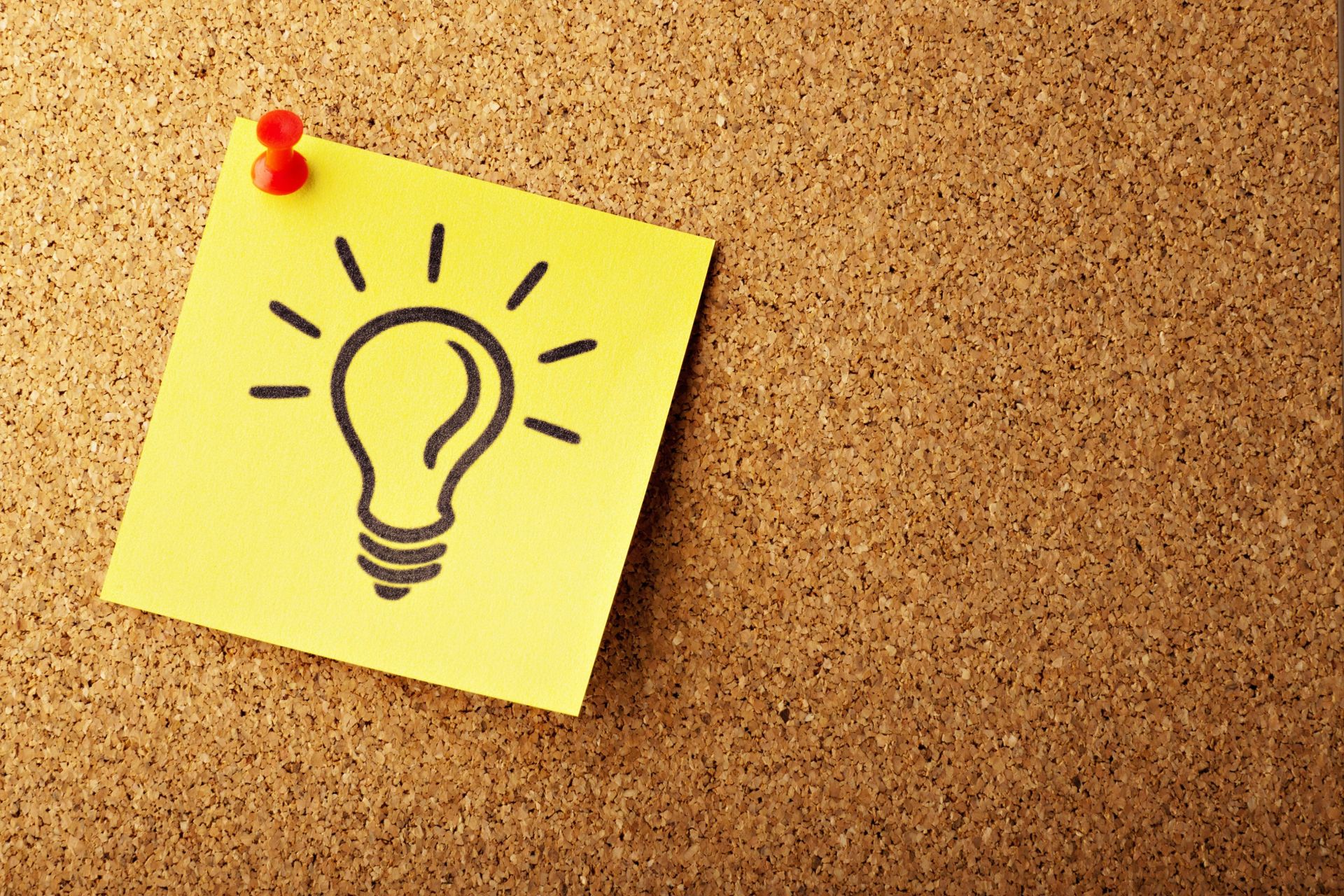 a yellow sticky note with a lightbulb drawing is pinned to a corkboard with a red pushpin