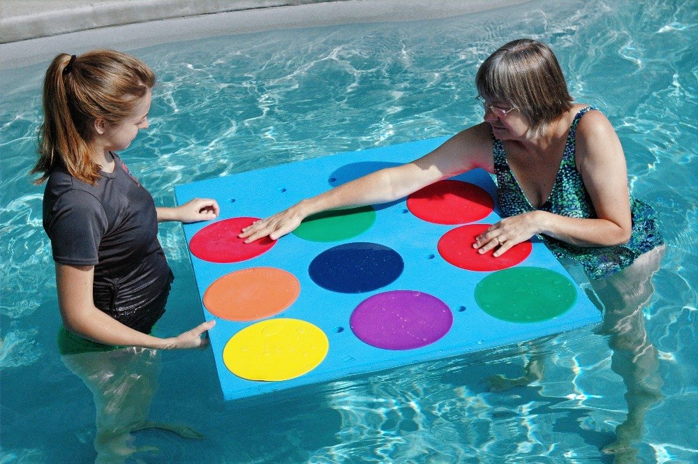 Two women in a pool work with a floating mat printed with colored circles.
