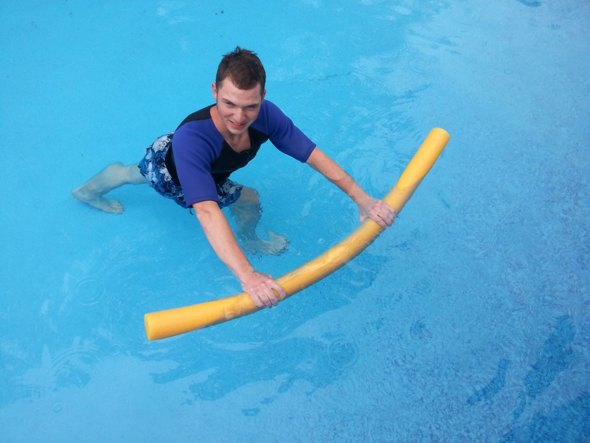 A smiling adult man stands in bright blue pool water and presses a yellow foam noodle down in front of him during an aquatic exercise