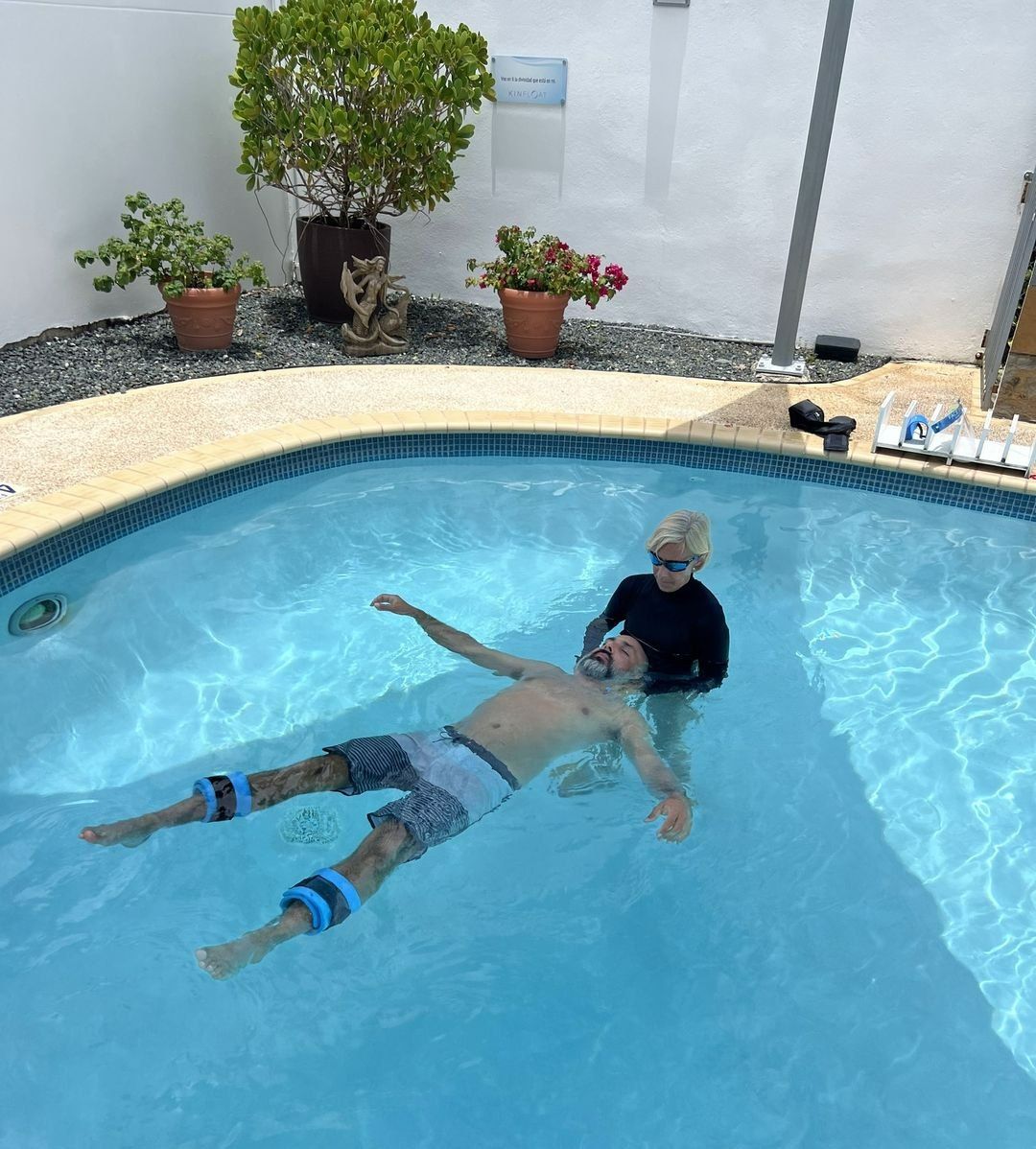 Two women in a pool play with a young child sitting on a red float with toy balls.