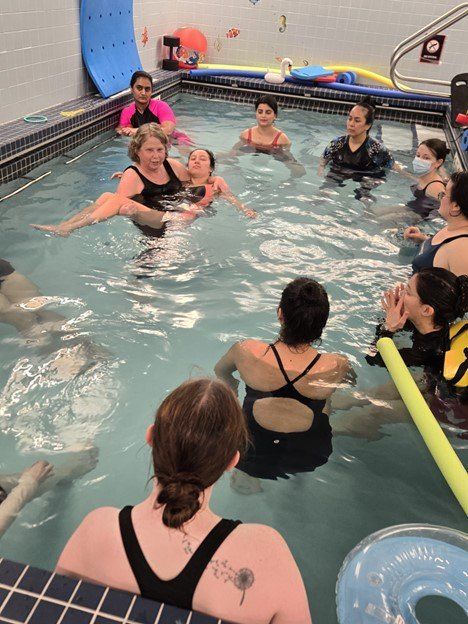 A group of people stands in an indoor pool, some holding foam noodles.