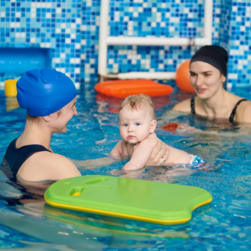 Two women in swim caps hold a baby in the pool beside a green kickboard.