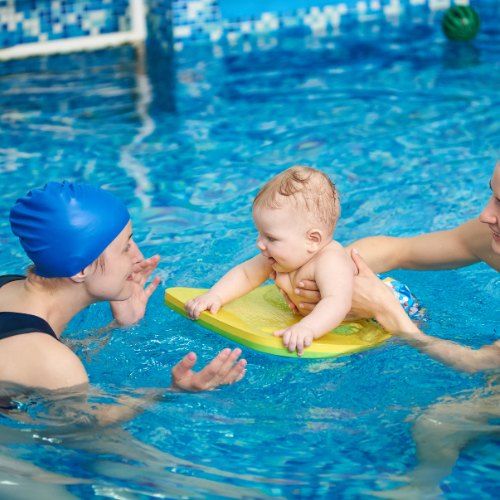 A baby lies on a yellow float in a pool while two adults in swim caps hold it.