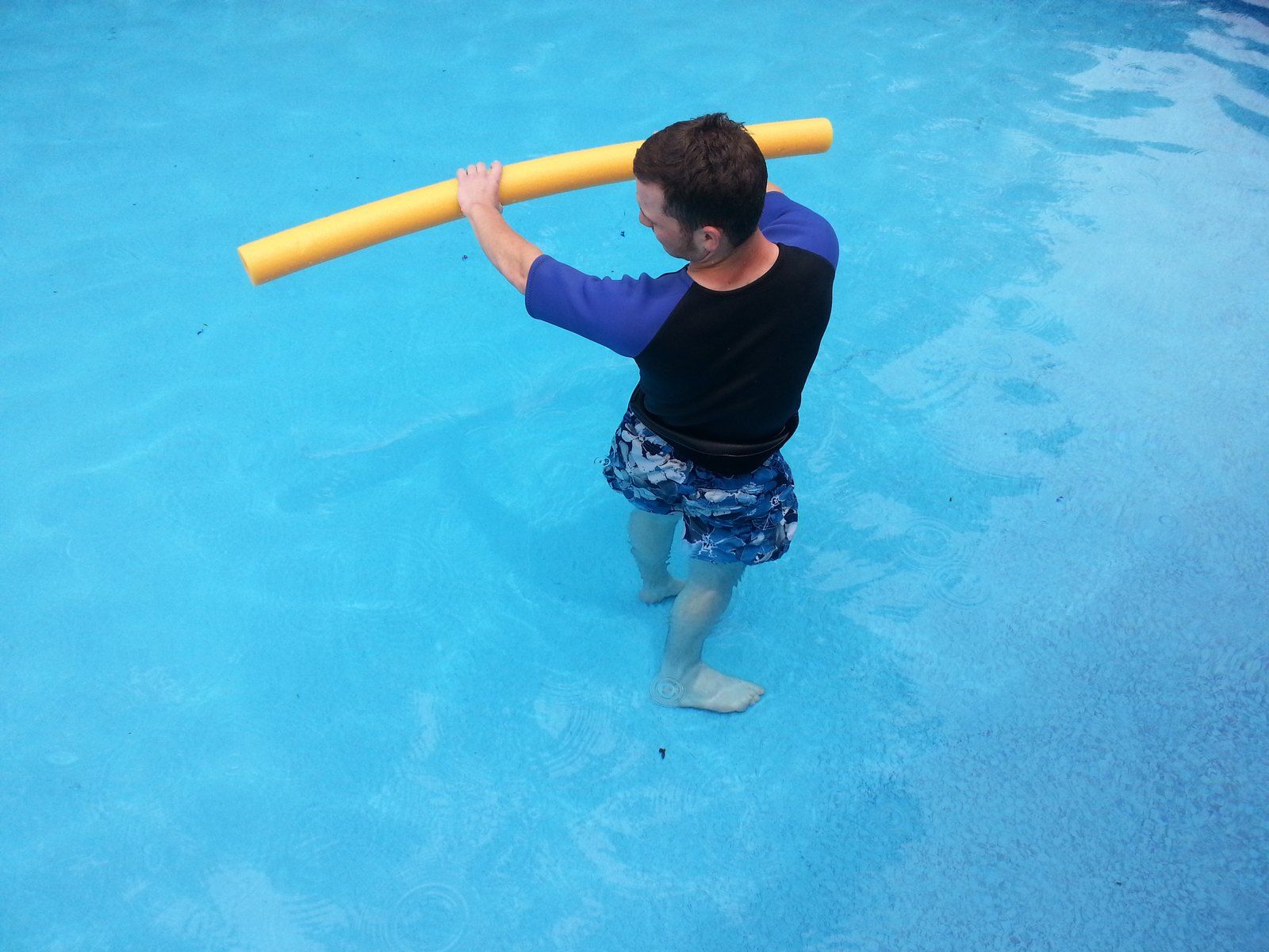 A man stands in a pool holding a yellow foam noodle overhead.