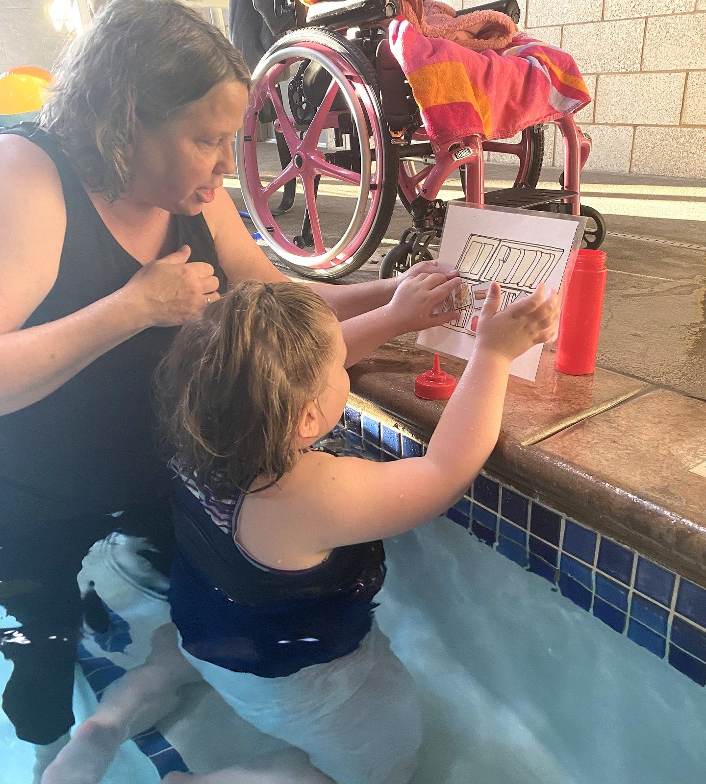 An ATU therapist and a young girl at the pool edge; the girl points to a picture card as they talk.