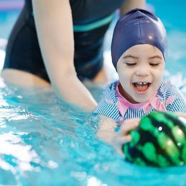 A smiling young child in a swim cap holds a striped ball in the pool, with an adult beside her.