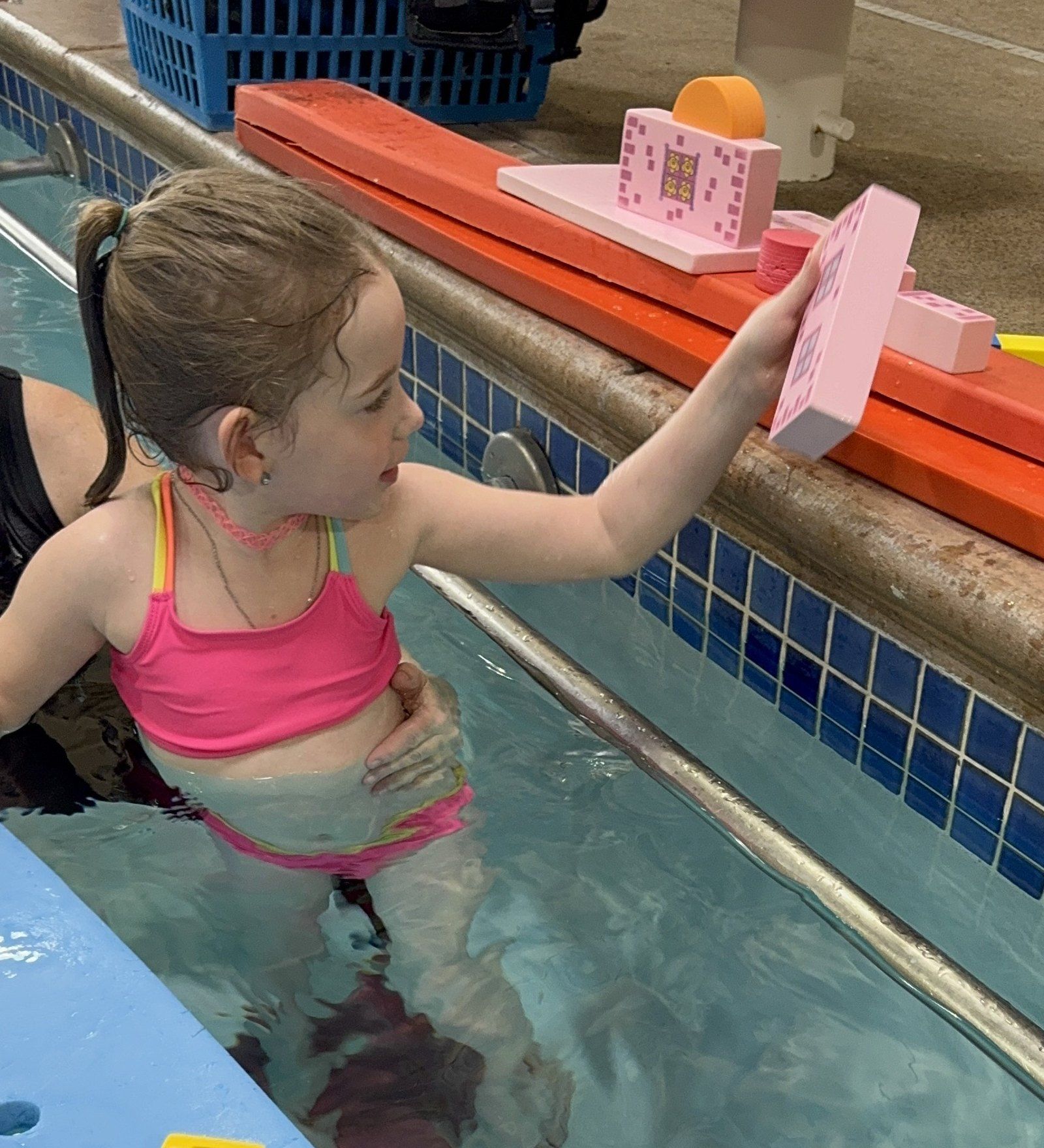 A girl at the pool wall reaches up to set a pink block on the deck.