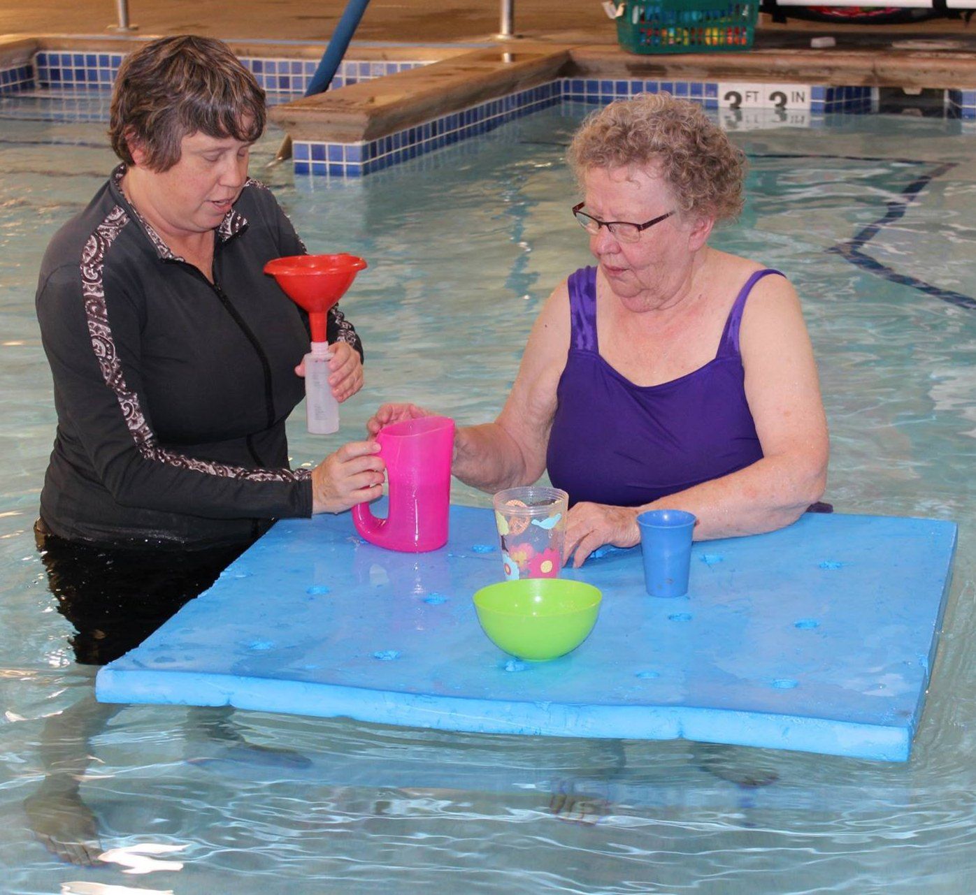Two women stand at a floating blue mat in a pool, pouring water between cups through a funnel.