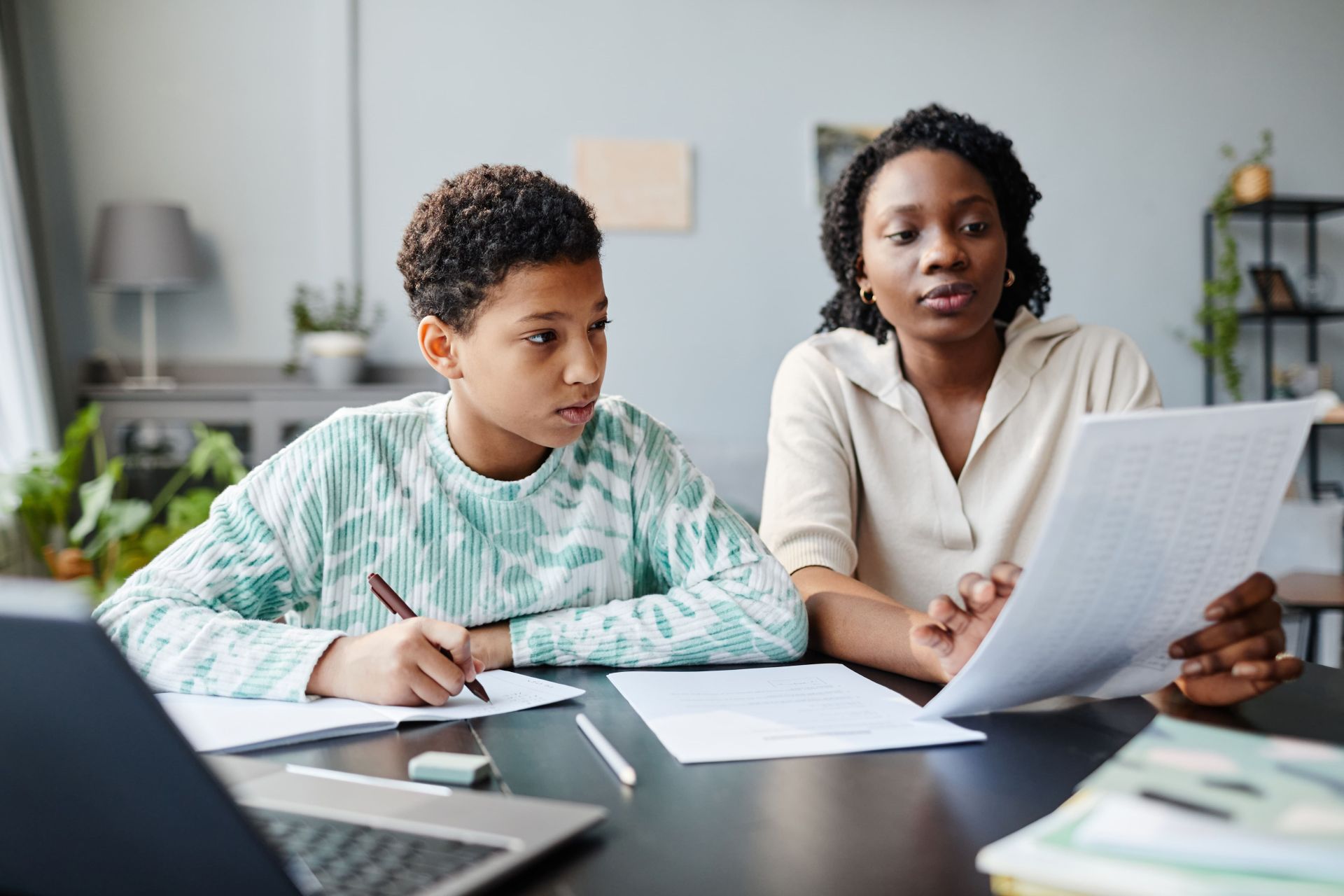 Two people sit at a table with papers, a laptop, and a pencil case, working together on homework in a cozy study room with shelves and plants.