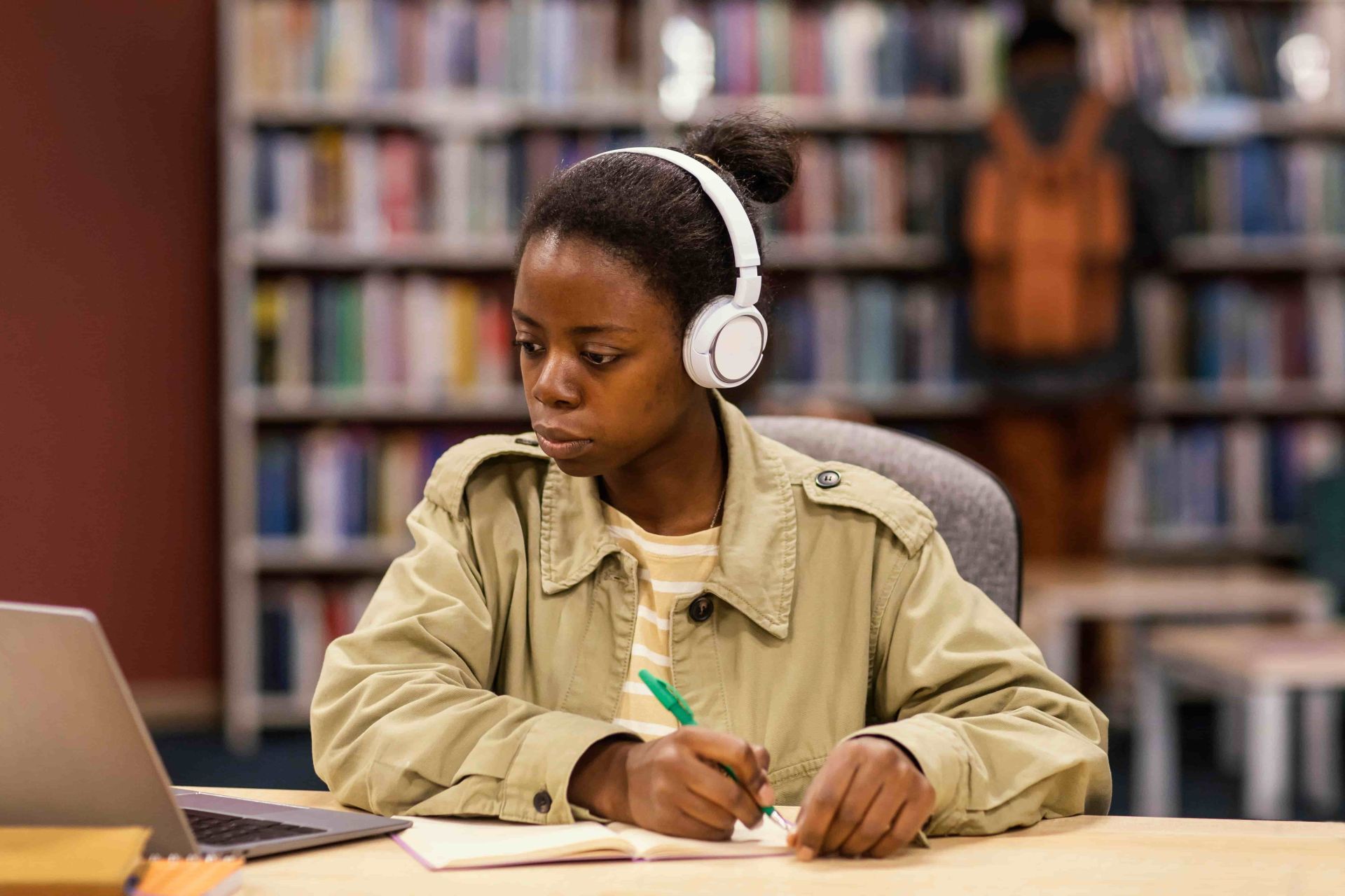 A student wearing white headphones and a light jacket sits at a desk in a library, studying with an open laptop and notebook while holding a green pen, with bookshelves in the background creating an academic atmosphere.