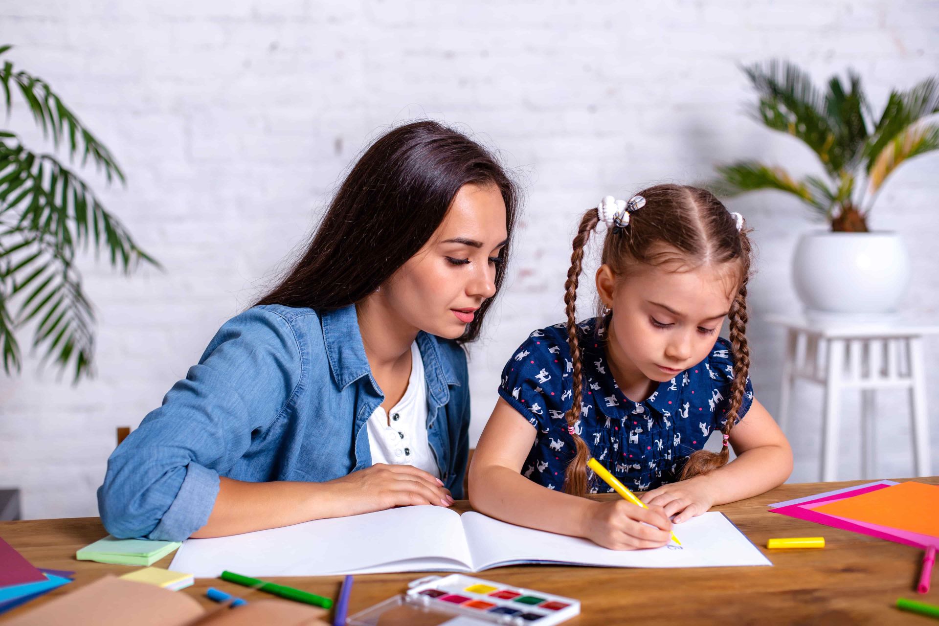 An adult and child sit at a wooden table with colorful pencils, markers, and a watercolor palette, as the child writes in a notebook with a yellow pencil, capturing a focused educational activity in a bright indoor setting.