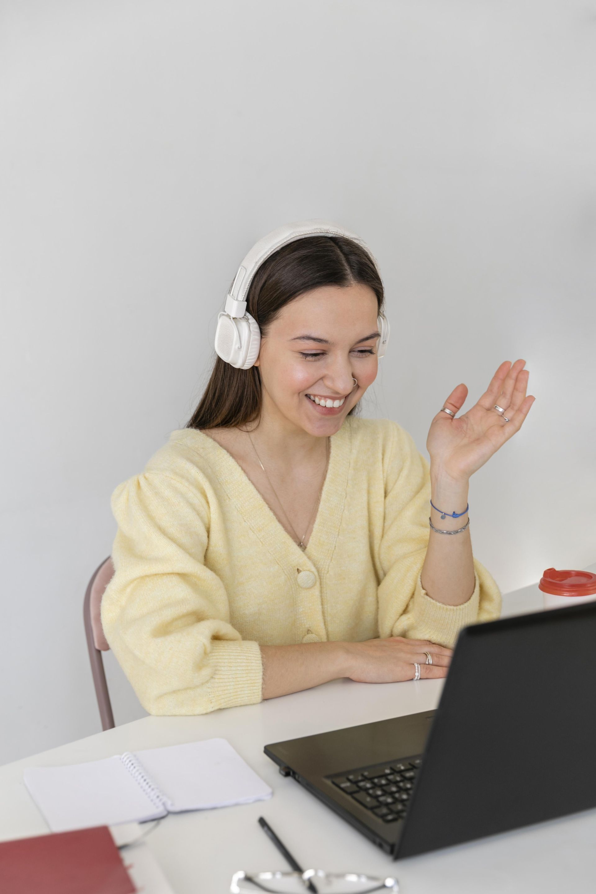 Smiling woman wearing headphones waves during an online video call while sitting at a desk with a laptop and notebook.