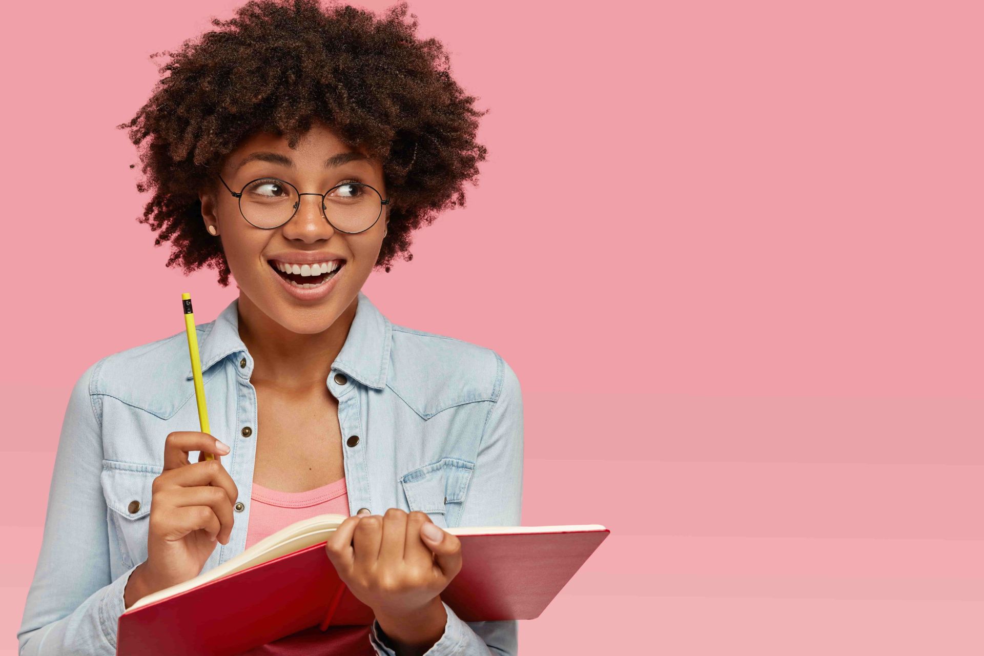 Student with curly hair wearing glasses and denim shirt, holding a red notebook and yellow pencil against a pink background