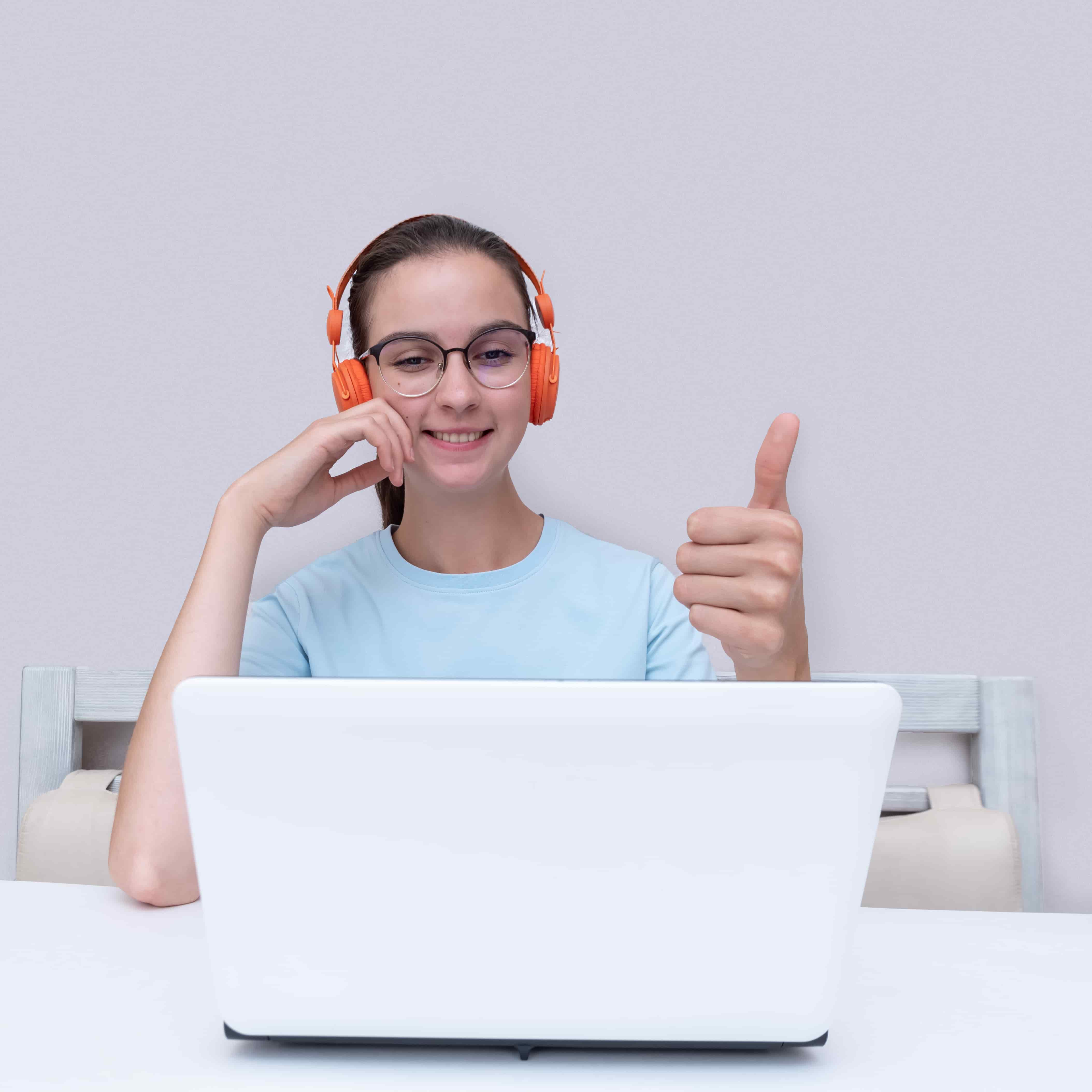 A happy teenage girl learning at home, wearing headphones and giving a thumbs-up while watching a video lesson on her laptop.
