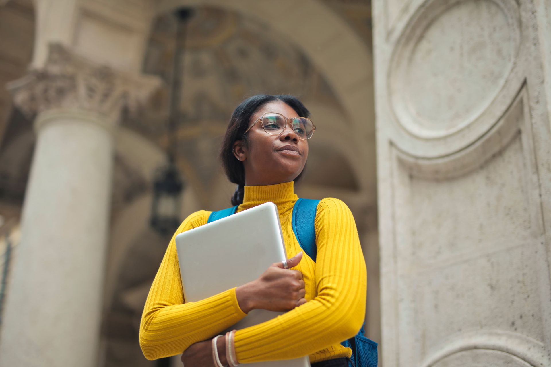 Student in yellow turtleneck with glasses and backpack holding laptop in front of stone columns and arched architecture.