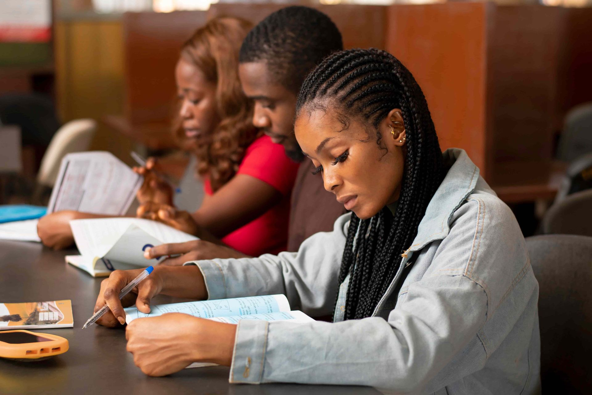 Three students studying at a library table, writing in notebooks and reading books with wooden partitions in the background.