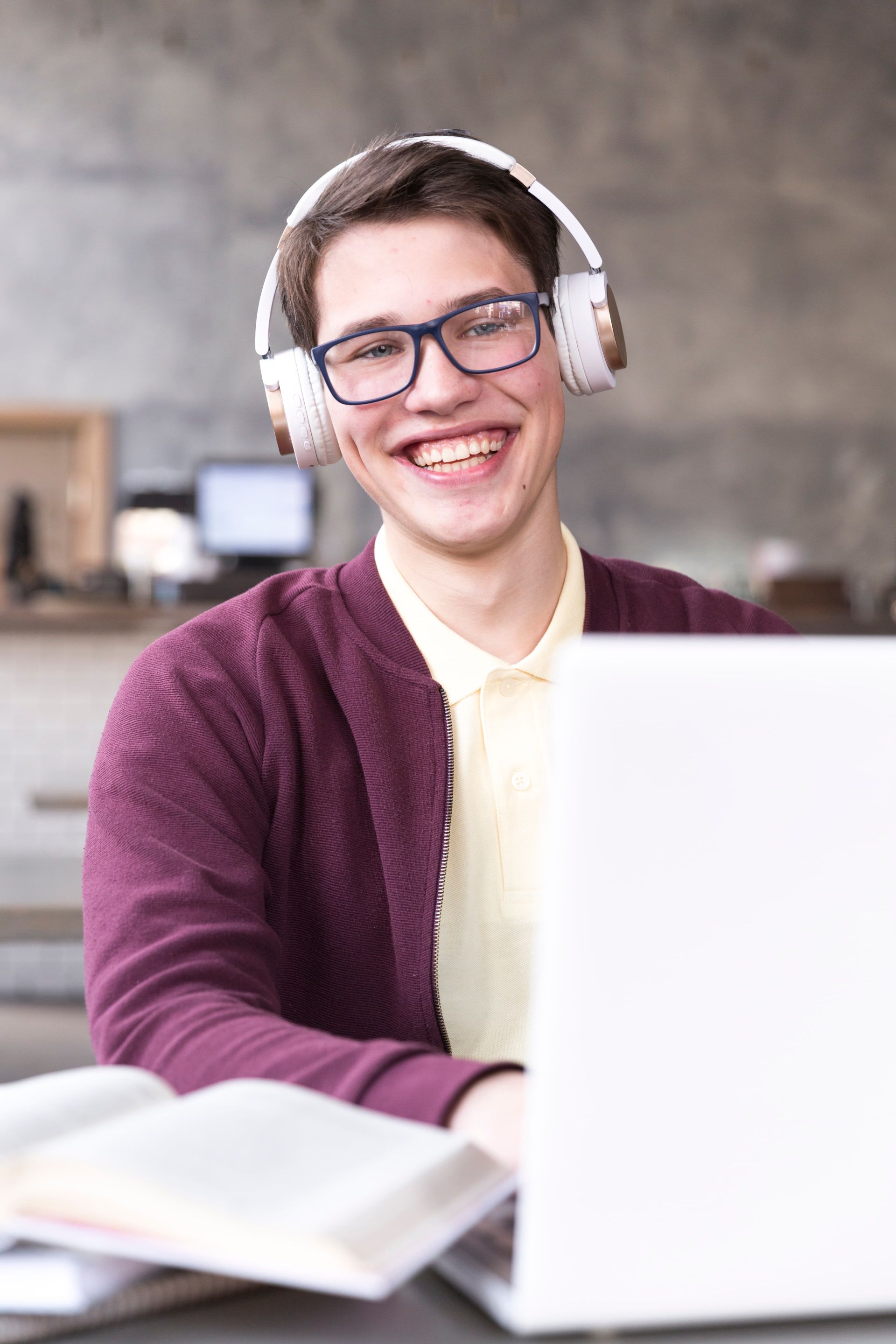 A smiling student wearing glasses and headphones working on a laptop with an open book on the table.