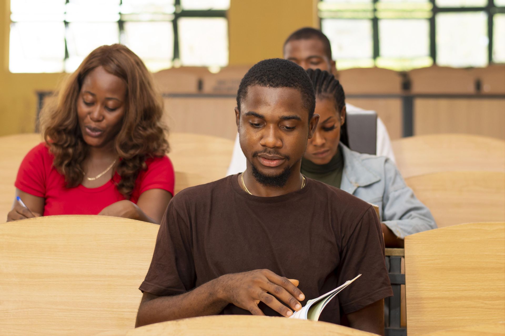Four students sit at wooden desks in a classroom with large windows, engaged in reading and writing, including one in a dark brown shirt looking at a book and another in a red shirt writing in a notebook, with two more students visible behind them.