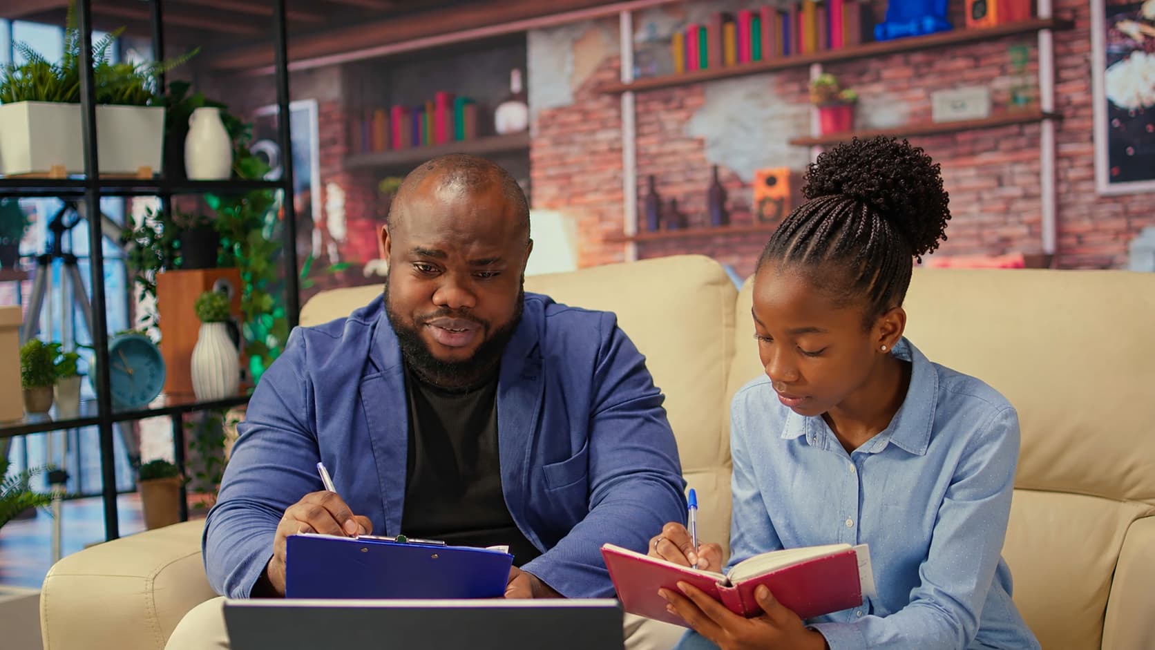 Two people sit on a beige couch with a laptop, clipboard, and notebook, working together in a cozy study space with shelves, plants, and books, depicting a tutoring or homework session.