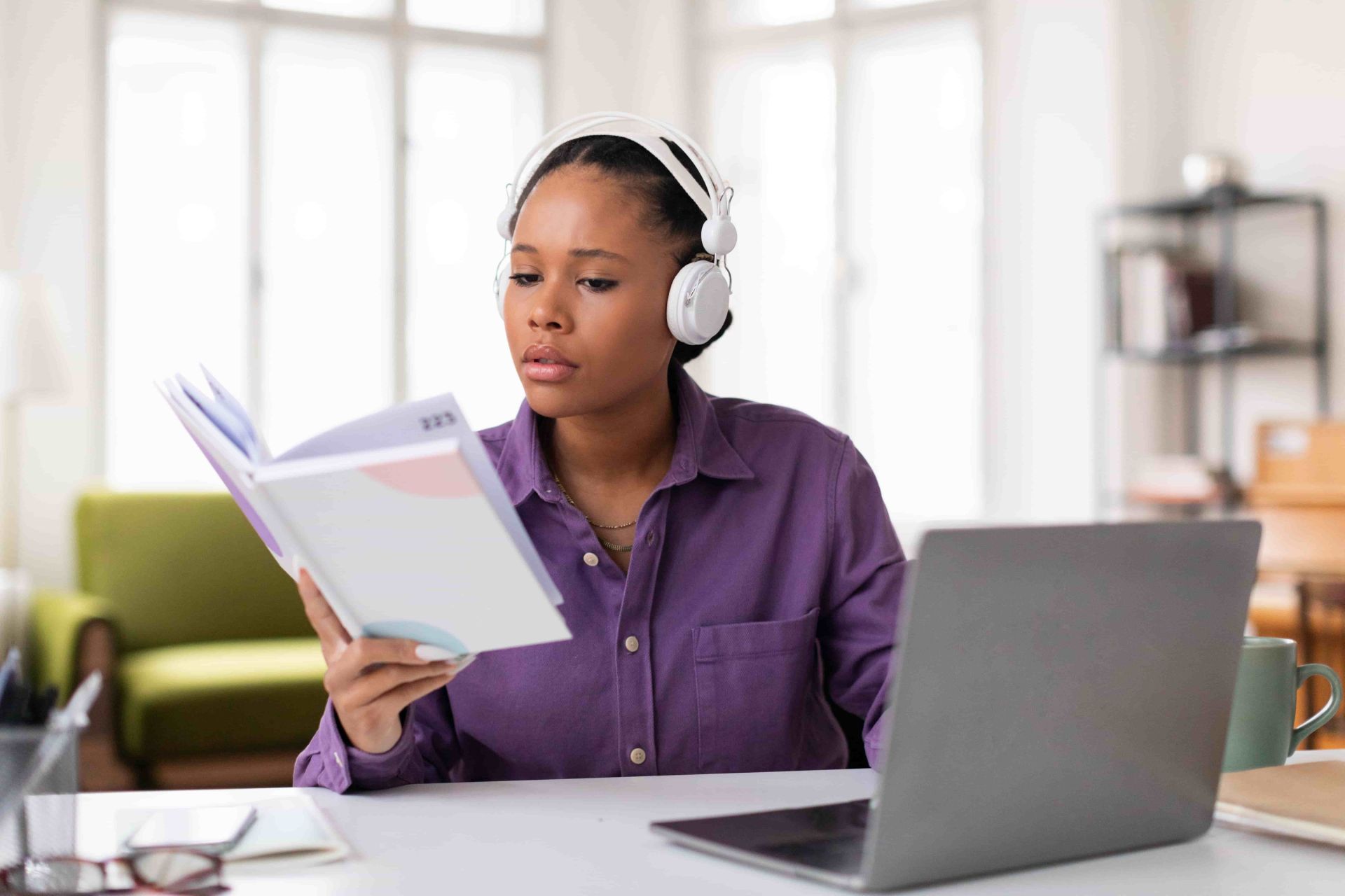 A student in a purple shirt sits at a desk wearing white headphones, reading a notebook with an open laptop, green mug, and eyeglasses nearby, in a bright study space with bookshelves and a sofa in the background.