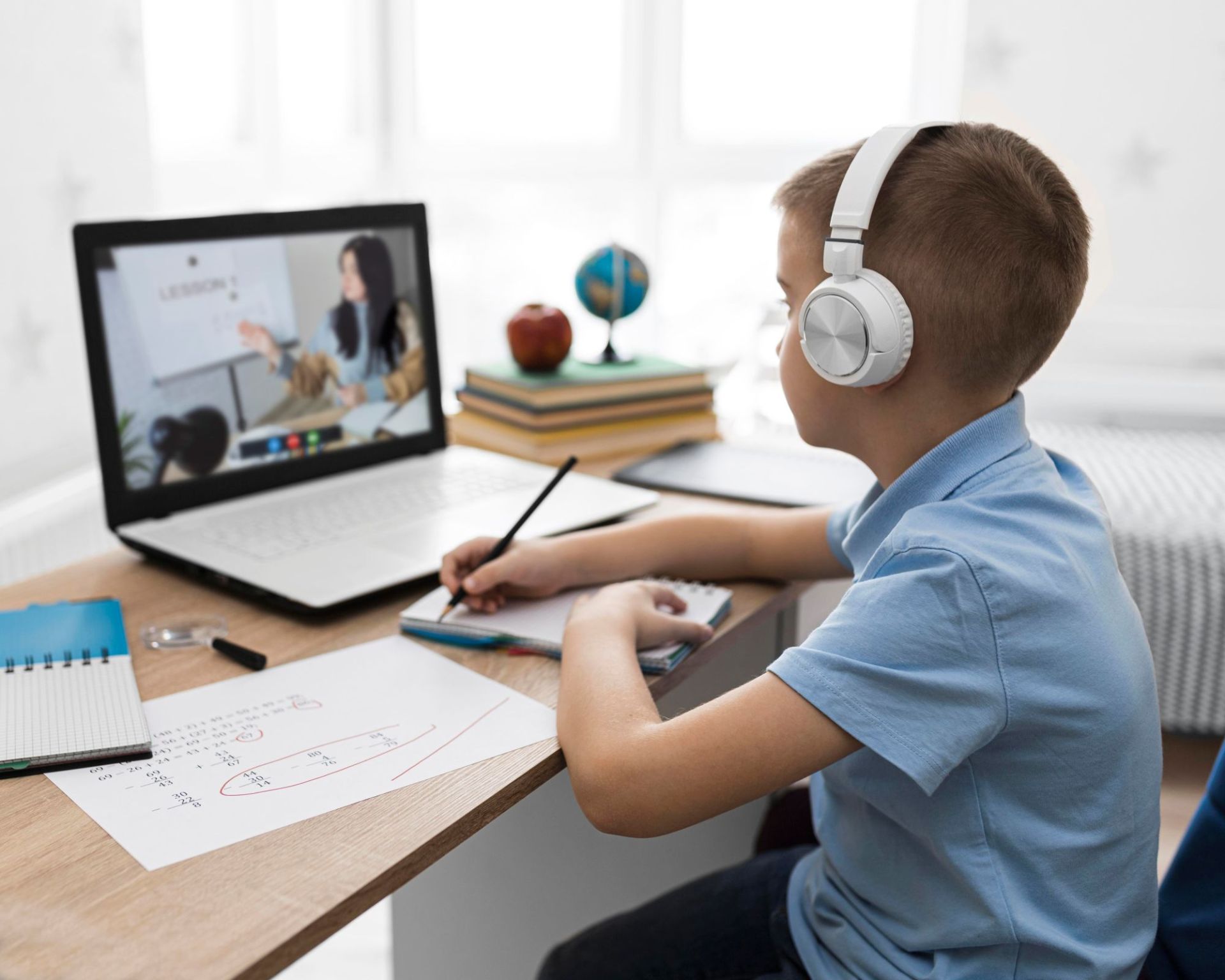 A young student wearing headphones sits at a desk, taking notes while attending an online lesson on a laptop. Books, a globe, and school materials are arranged around the workspace.