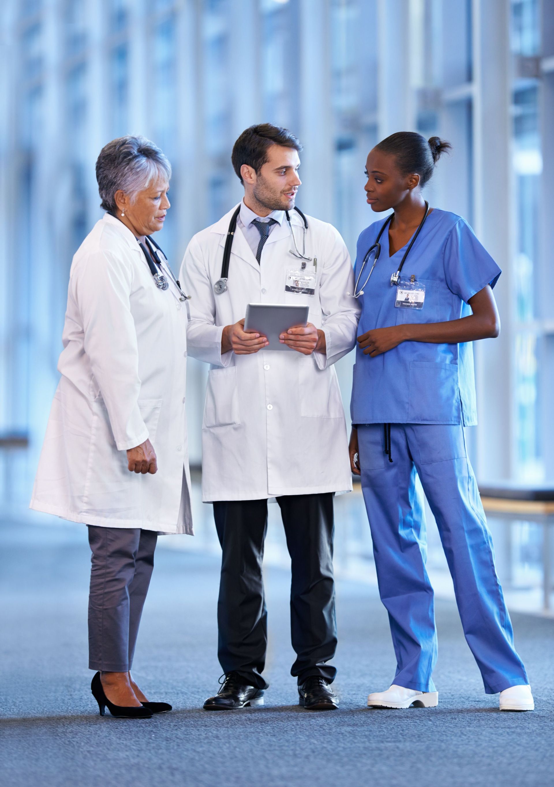 Three healthcare professionals stand together in a hospital corridor, wearing medical uniforms and discussing information displayed on a digital tablet.