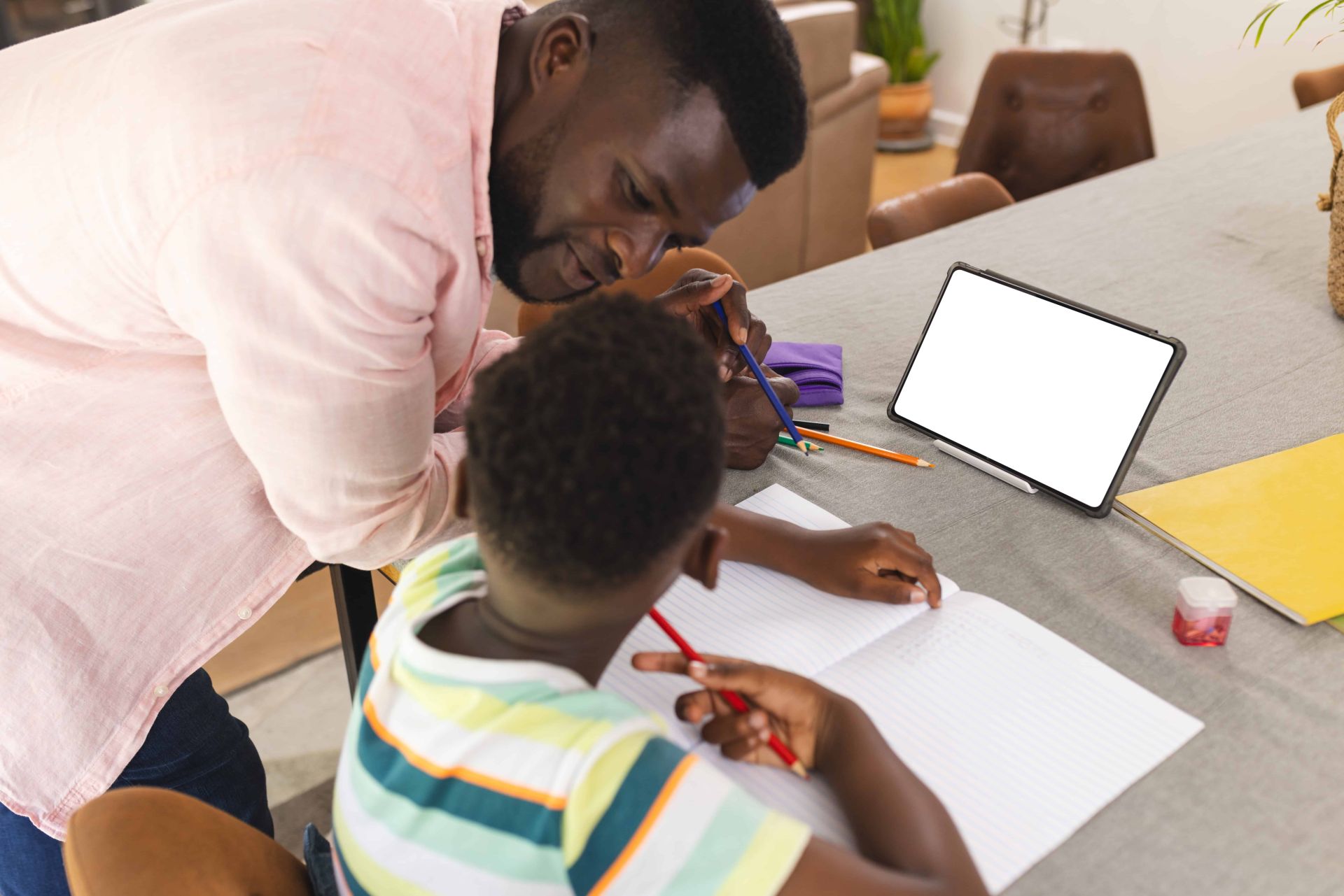Two people at a table engaged in a tutoring session, with one assisting the other who is writing in a notebook, surrounded by coloured pencils, a tablet, and study materials in a casual home learning environment.