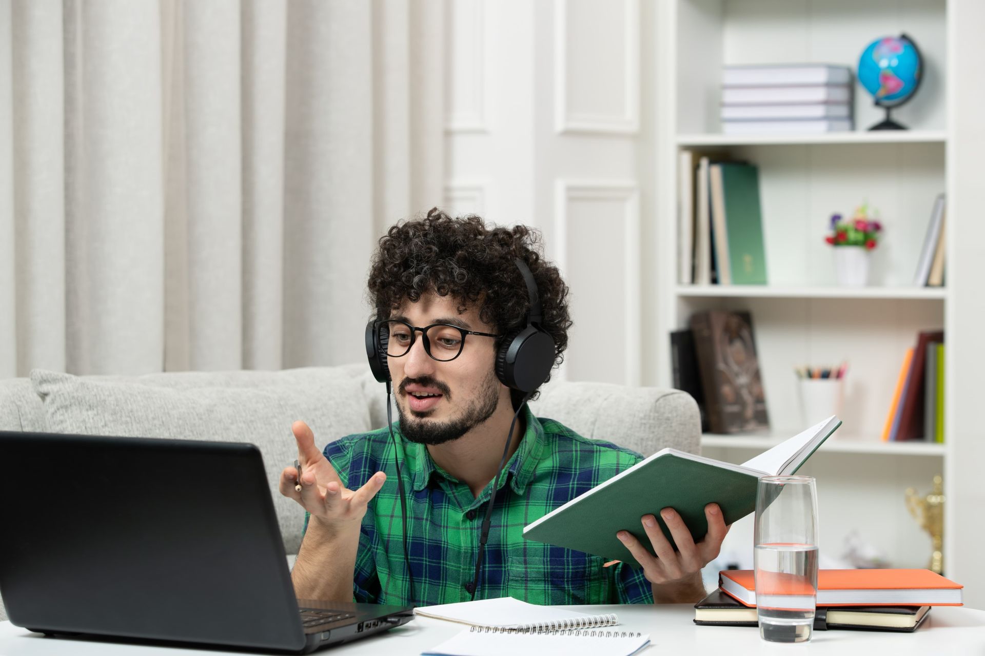 A student wearing headphones and glasses sits on a sofa with a green book, gesturing while looking at a laptop during an online study session, with notebooks, a closed orange book, and a glass of water on the table.