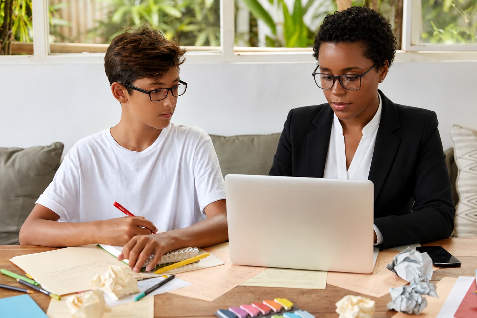Two individuals sit at a table with a laptop, notebooks, pens, and papers, actively engaged in a tutoring session in a bright indoor space with large windows and greenery outside.