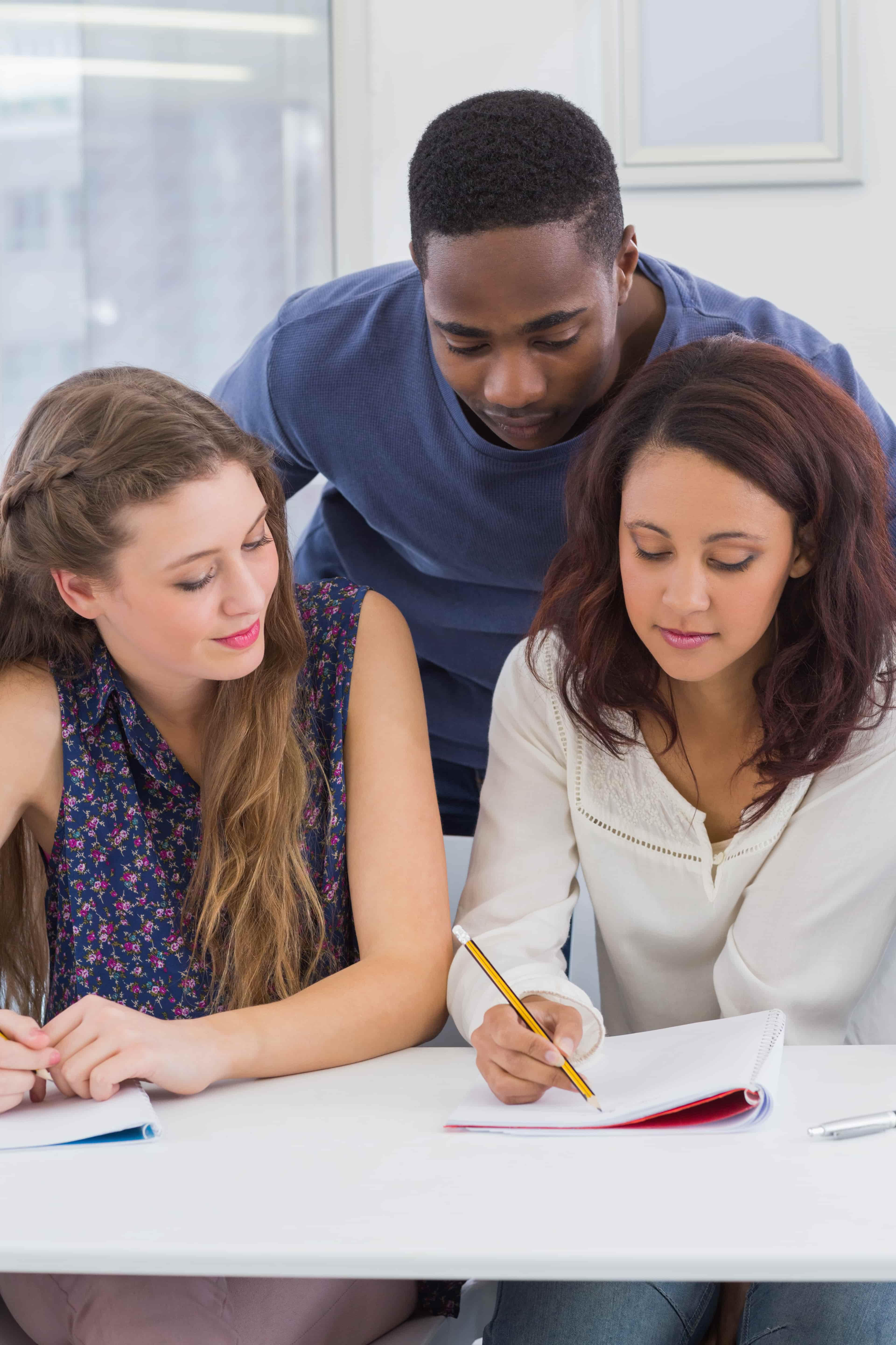 Three individuals gather around a white table in a bright classroom‑style setting, with two seated writing in notebooks and one standing behind them looking on, capturing a collaborative study or homework activity with large windows in the background.