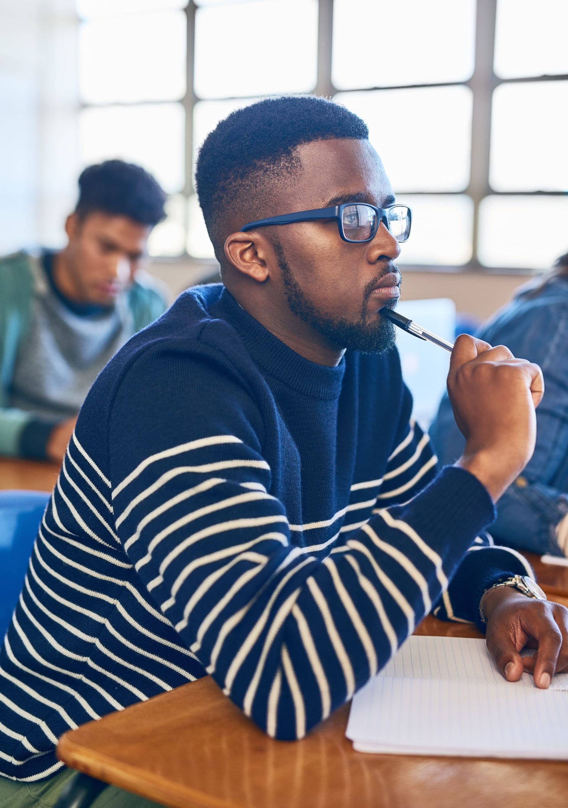 Student in classroom wearing striped sweater and glasses, holding pen thoughtfully with open notebook on desk