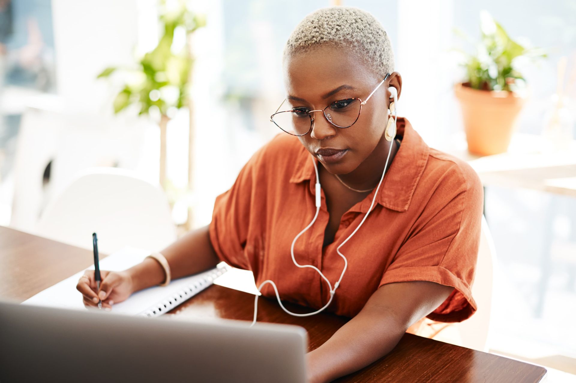 A student in an orange shirt wearing glasses and earphones writes in a notebook while looking at a laptop, with potted plants and natural light in the background, creating a modern and focused study environment.