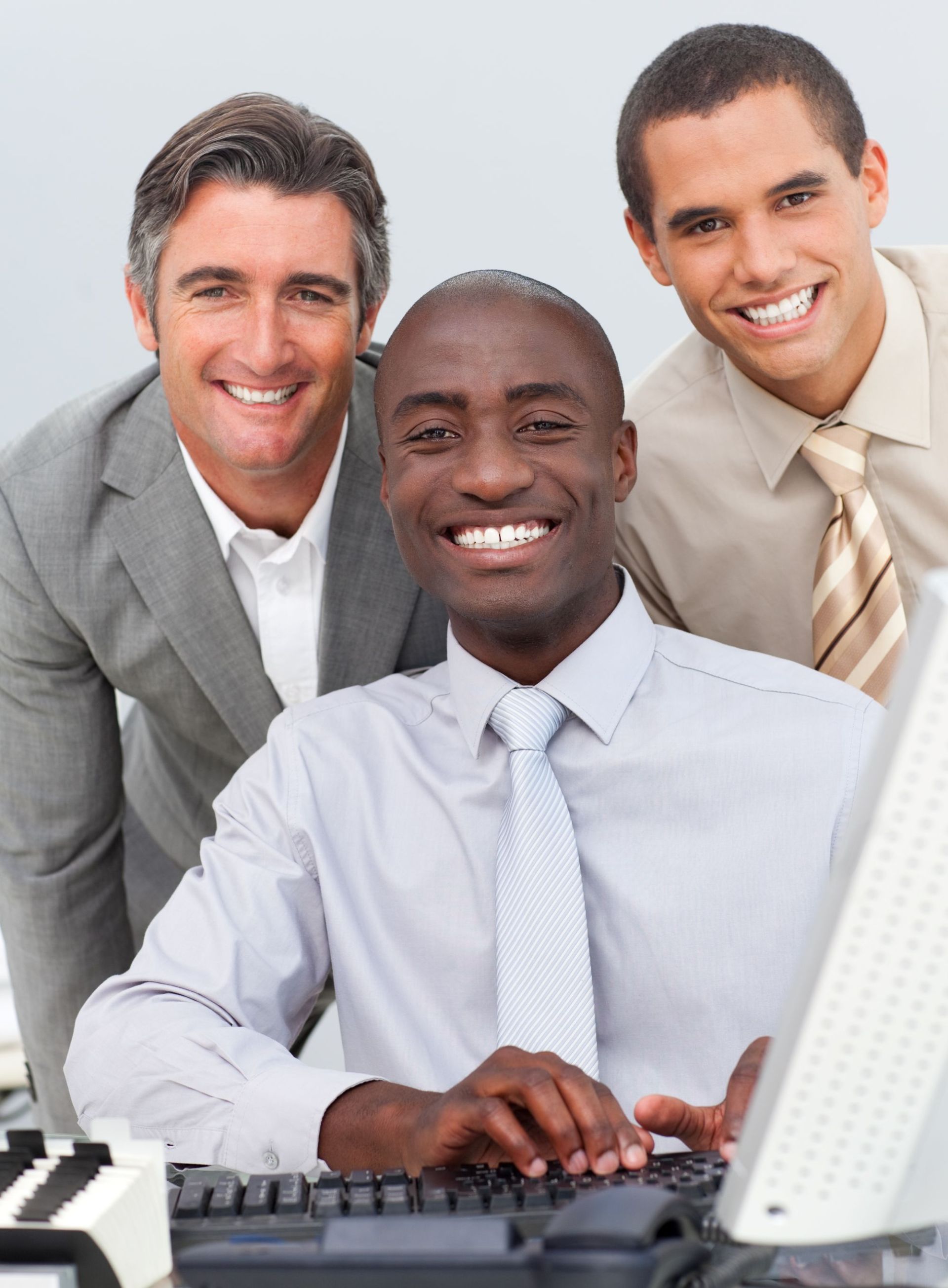 Three colleagues in business attire gather around a computer workstation, with one person seated typing on a keyboard while the other two stand behind observing the screen.