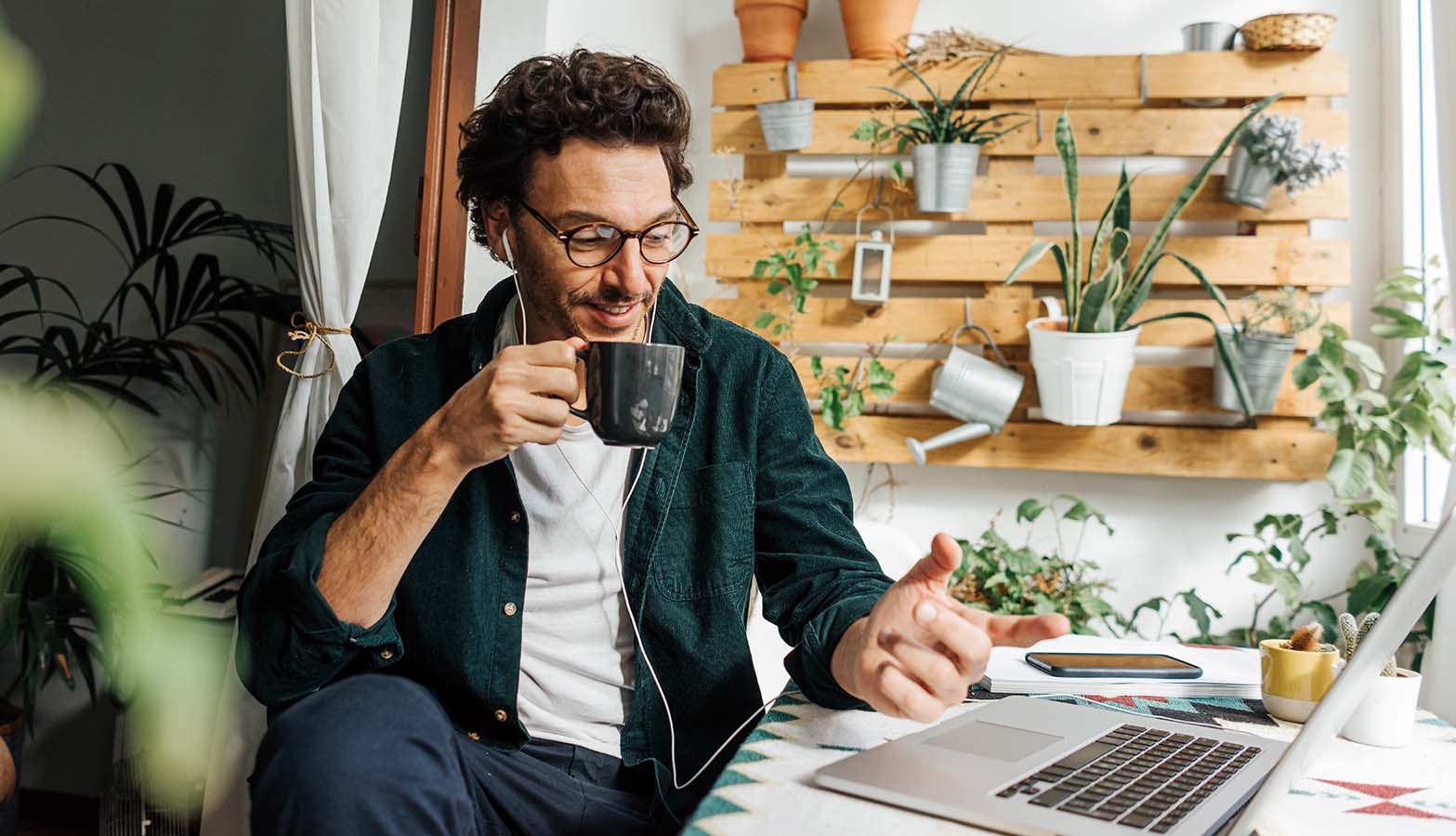man sitting at desk at home with plants behind him while taking a course online and drinking his coffee
