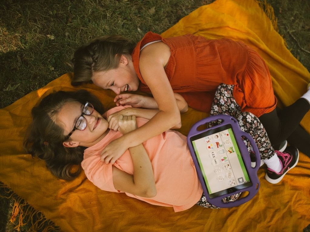 two 8 year old girls laughing on a picnic blanket one has assistive technology