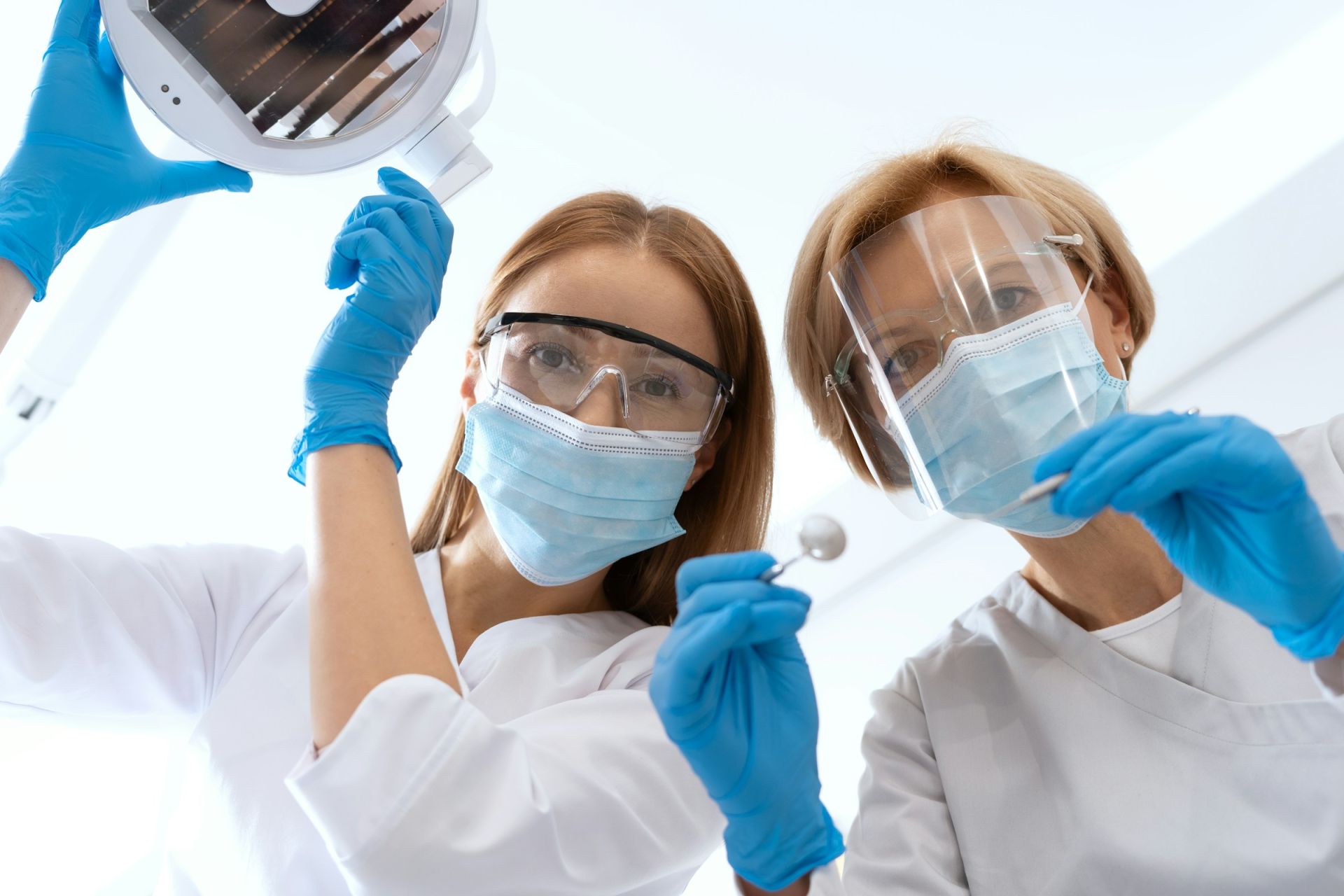 Two dental providers smiling behind masks and adjusting a dental light, set from the view of the website user as the patient looking up at them