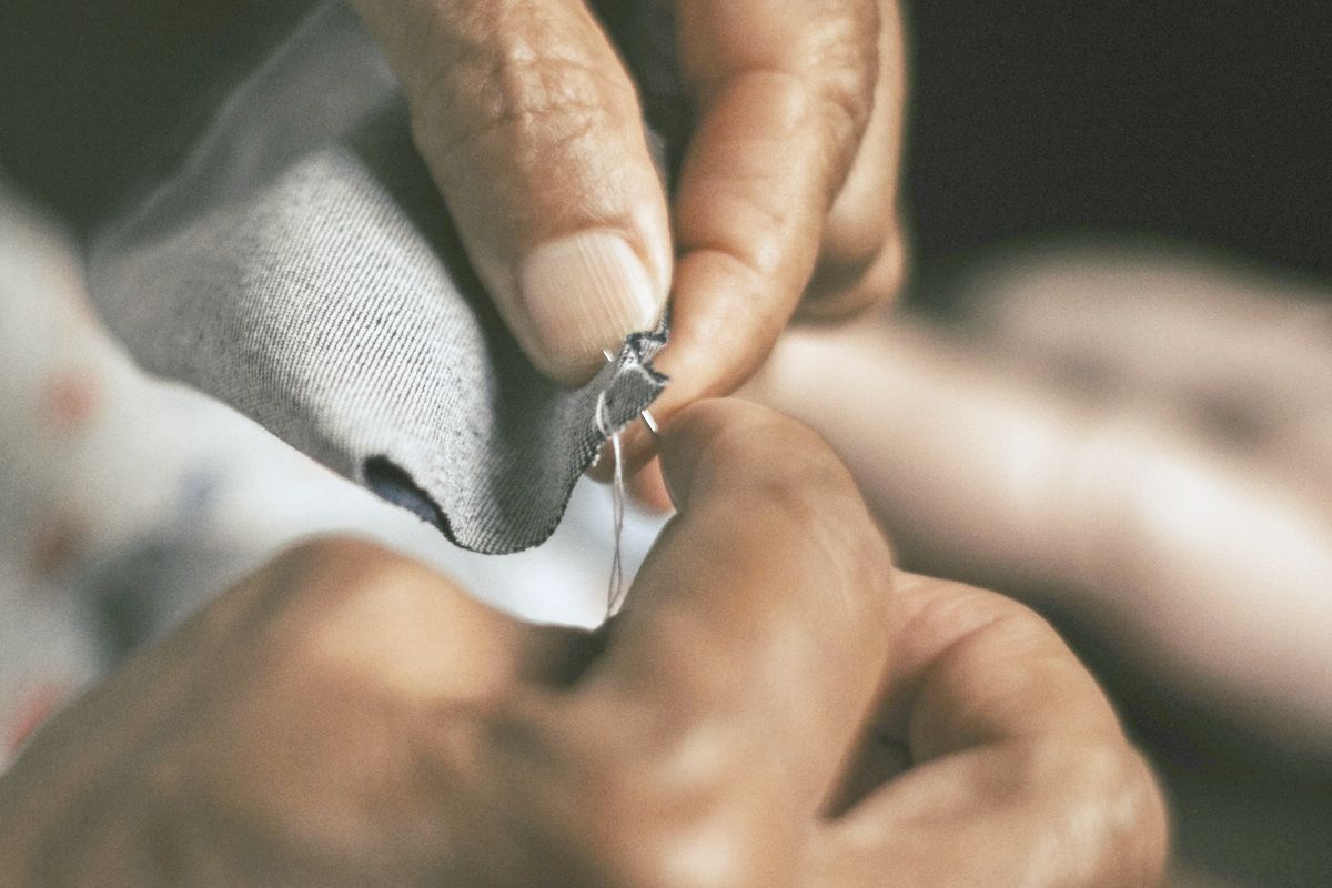 Two hands stitching a hole closed in grey cloth, the needle mid-stitch