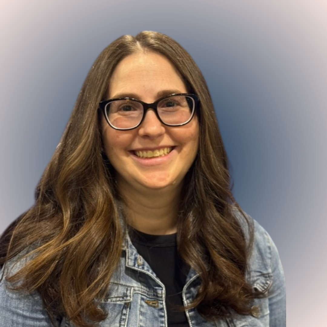 Image of a woman standing with arms crossed, smiling at the camera Image of a woman, Moira Bushell, smiling with brown hair and glasses in front of a tree