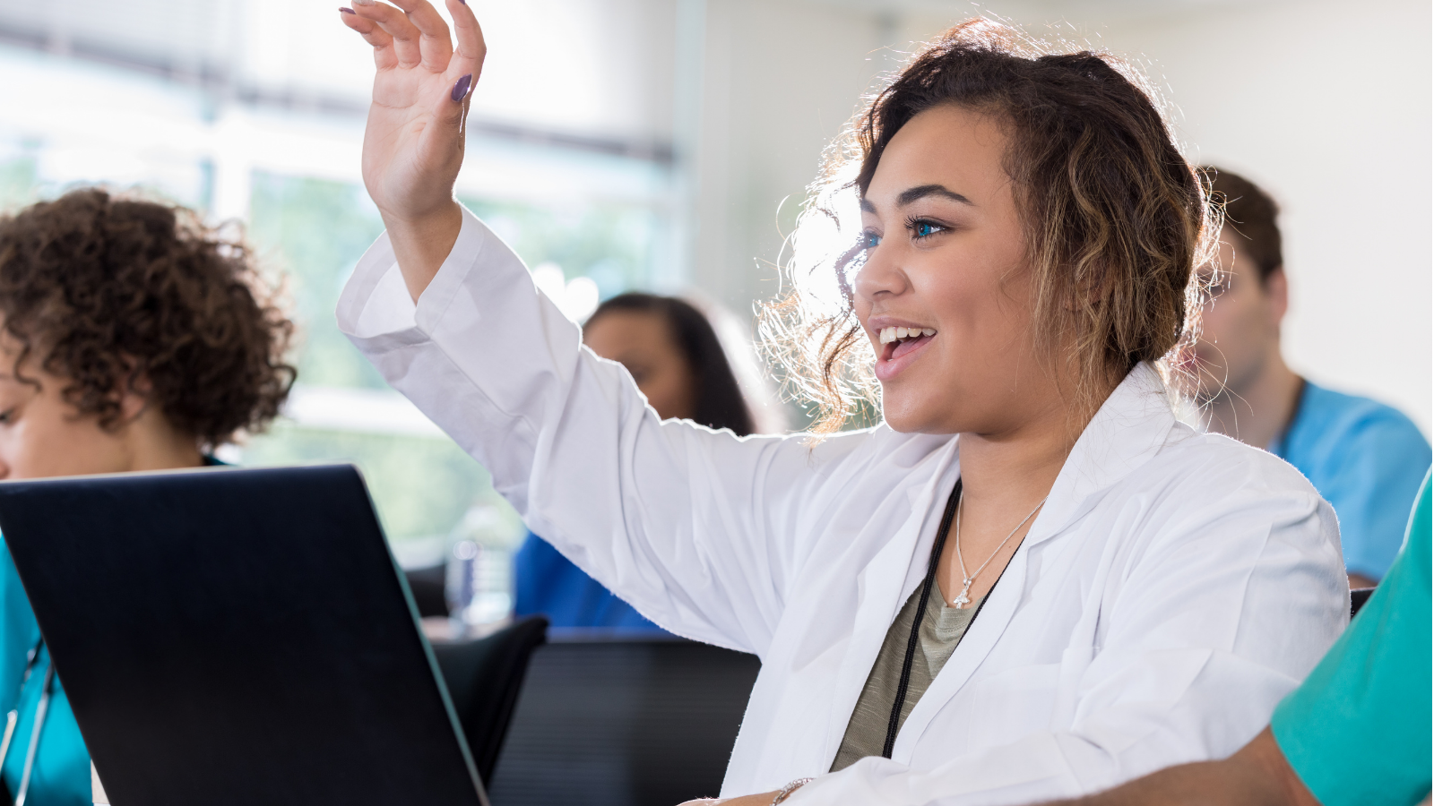 female with dark skin and hair in white lab coat sitting at a table raising her hand