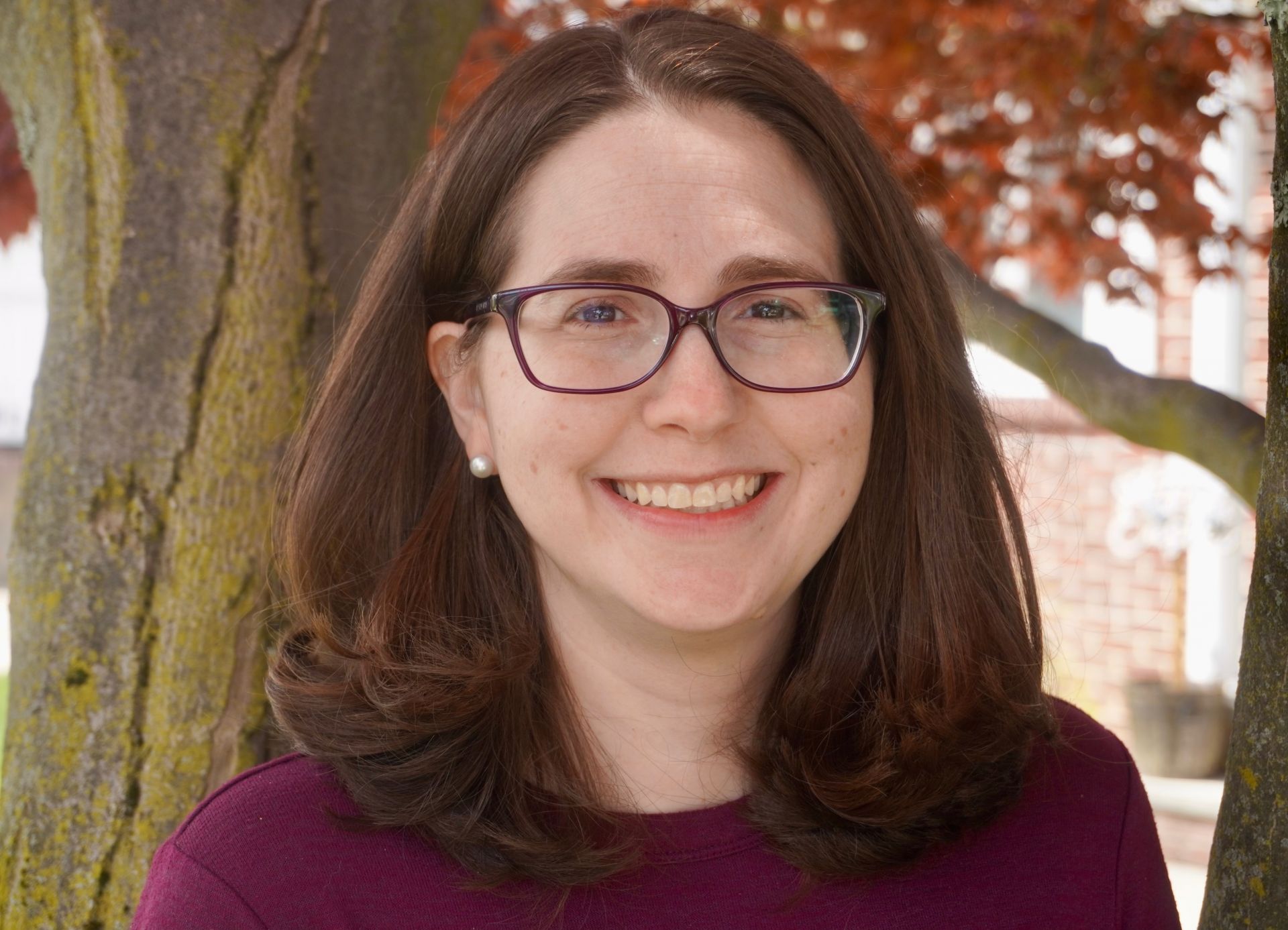 photo of a woman smiling with brown hair and glasses in front of a natural background