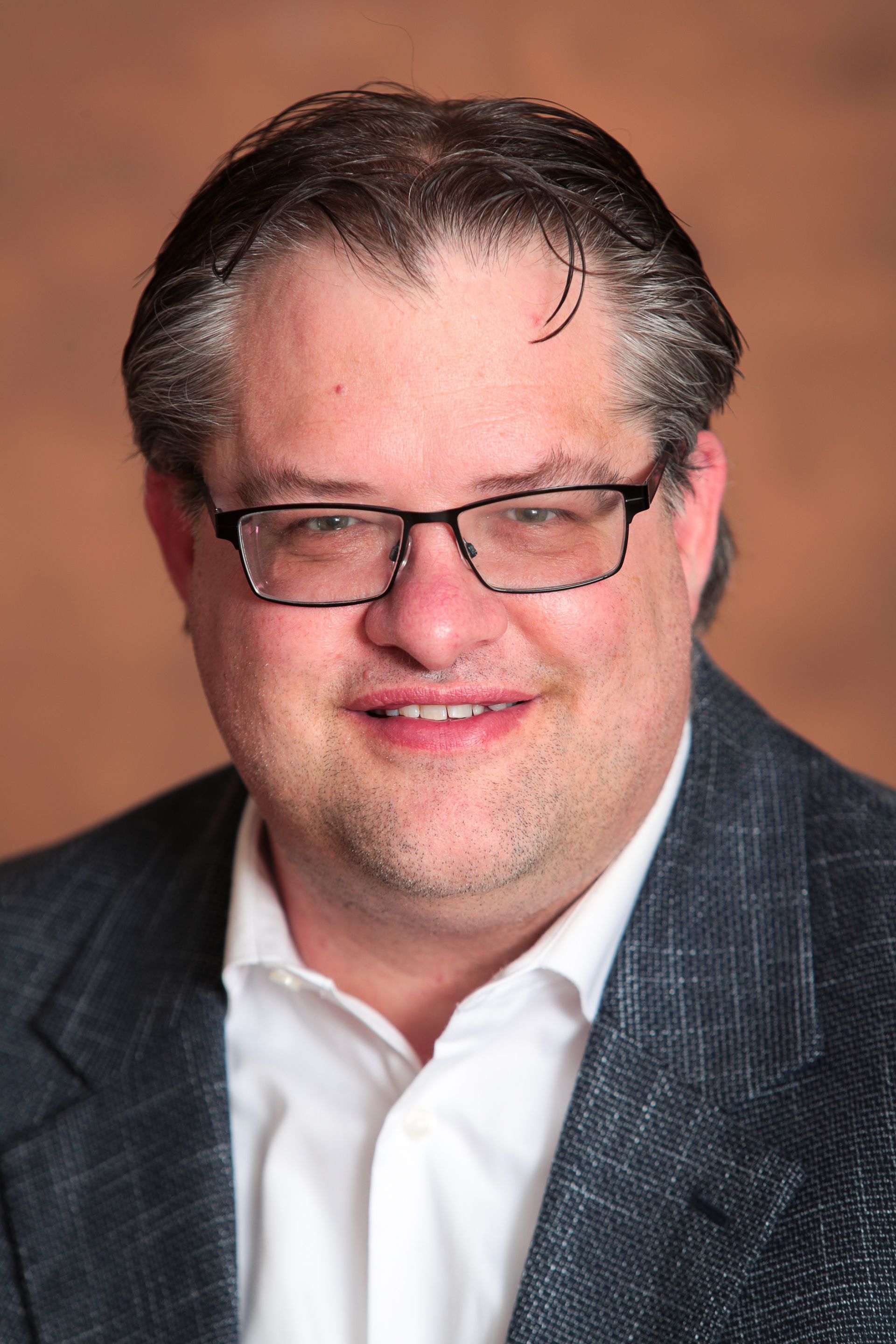 Image of a smiling white man with glasses in a suit jacket with white shirt in a formal portrait background 