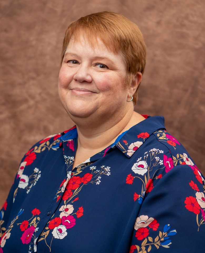 a smiling woman with short red hair, light skin, with a blue shirt with red floral and white pattern.