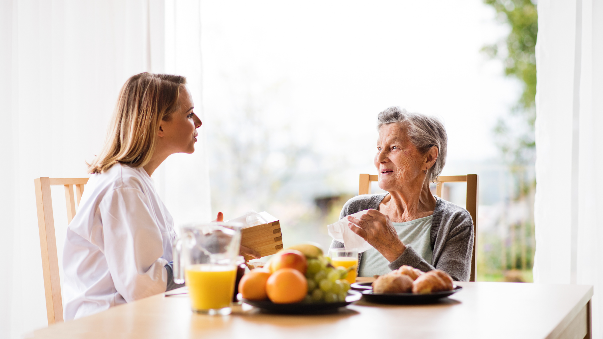 Photo of a young woman in a white lab coat sitting a table with an older woman , both drinking from cups