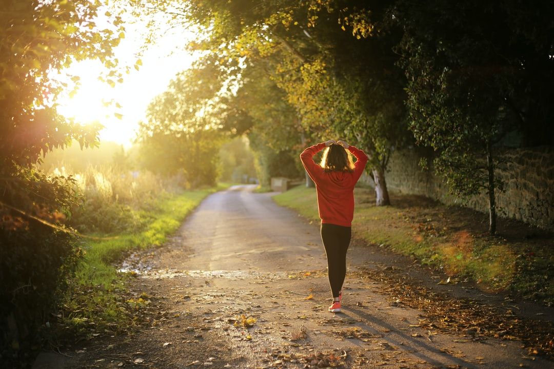 photo of back of woman walking a road with trees and grass and the sun shine through the trees