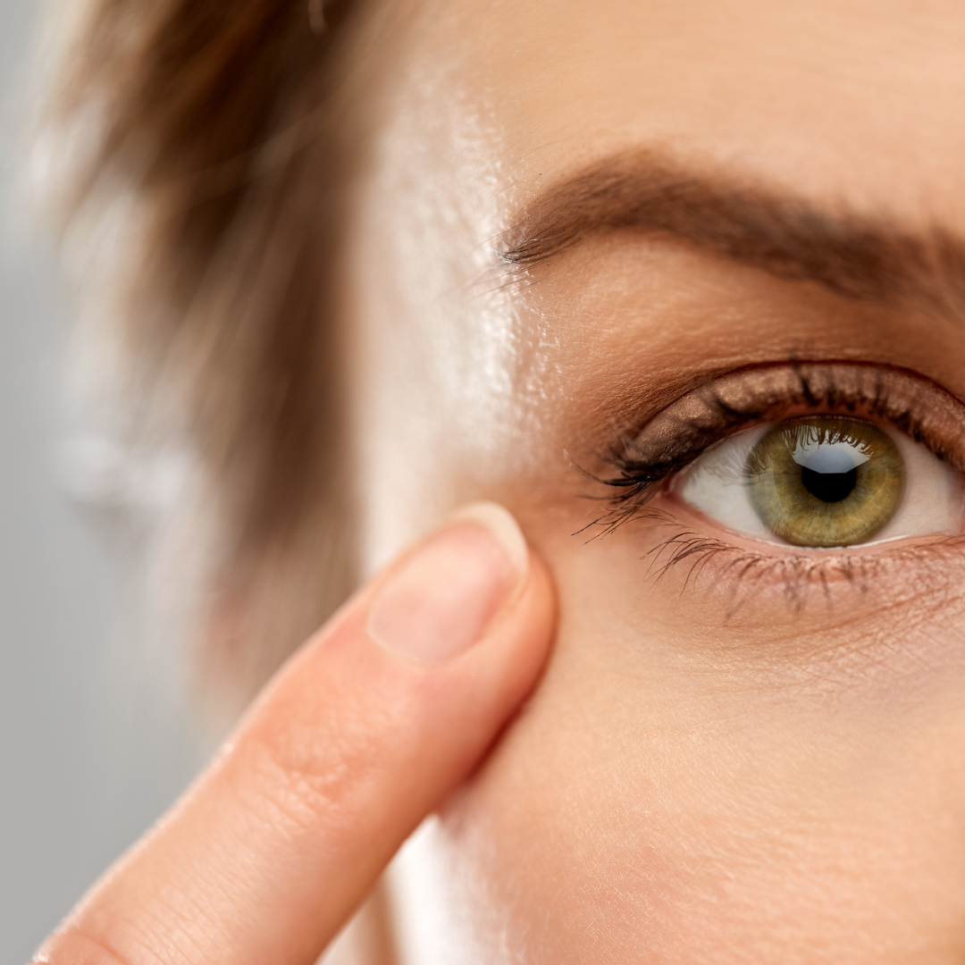 image: of a woman's eye, the eye is green, skin is light, eyebrows and hair are brown.  There is a finger without nail polish pointing to the eye