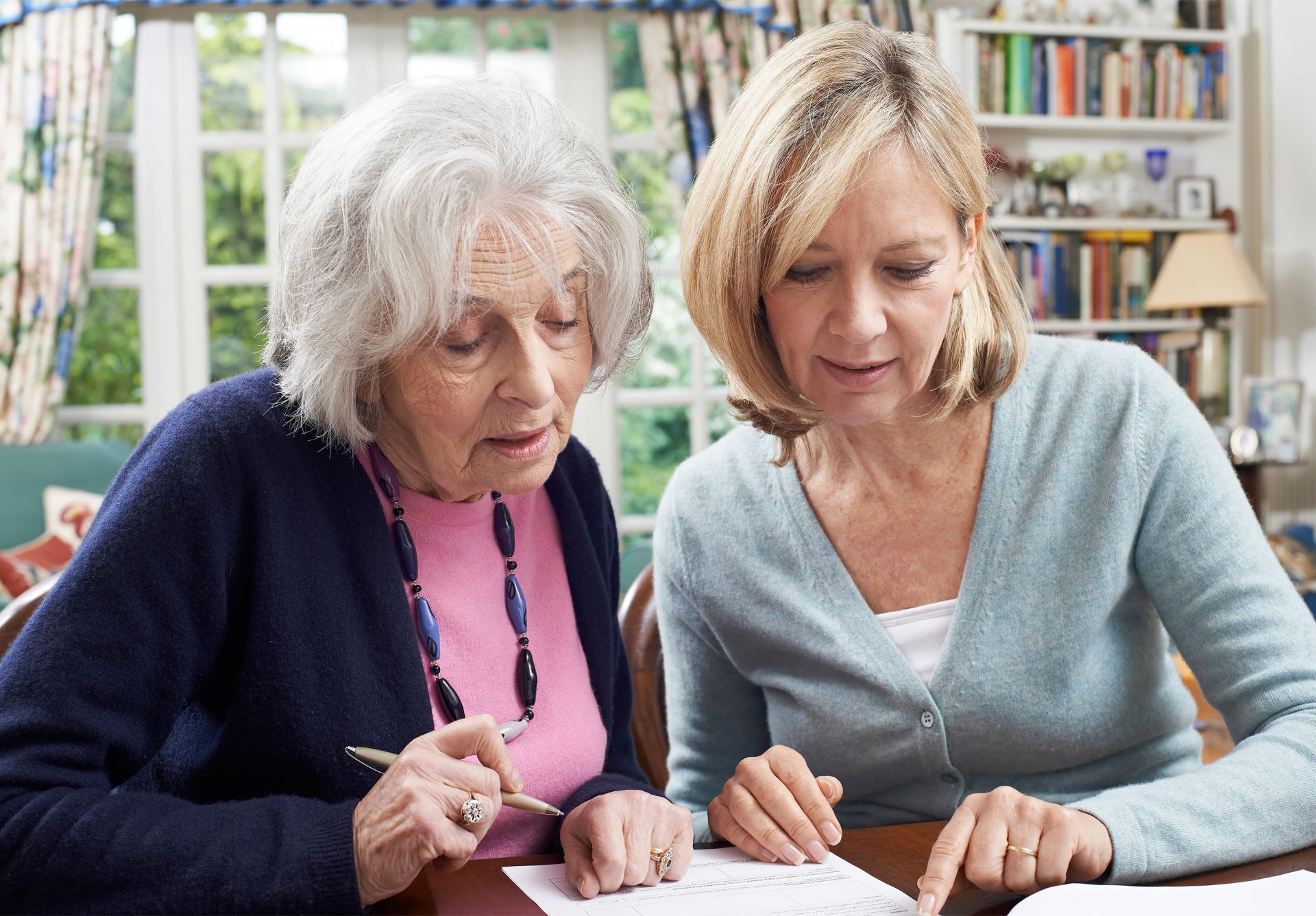 Older woman holding pen, middle aged woman assisting her at table