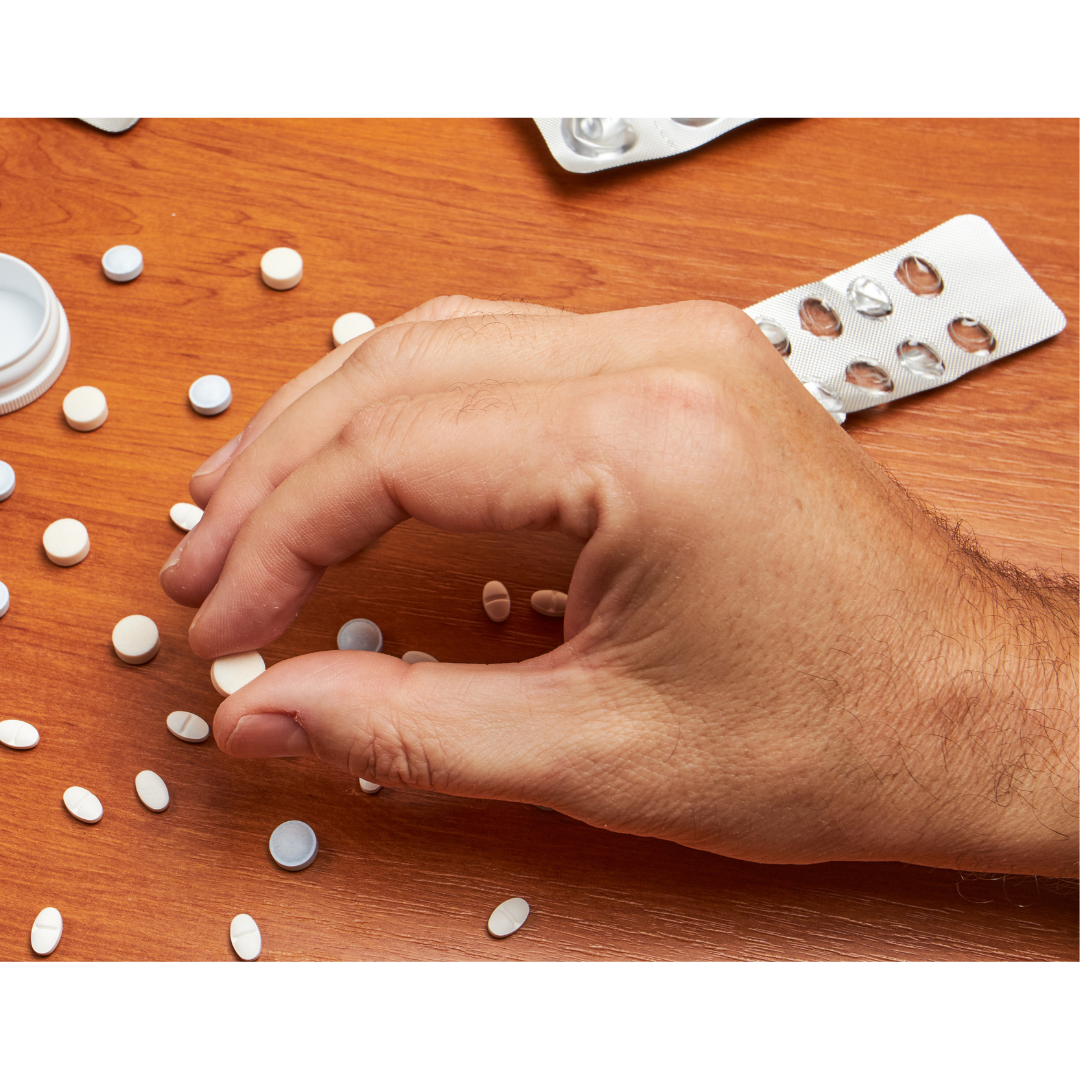 brown table with white pills on it, one hand with light skin picking up one pill between index finger and thumb