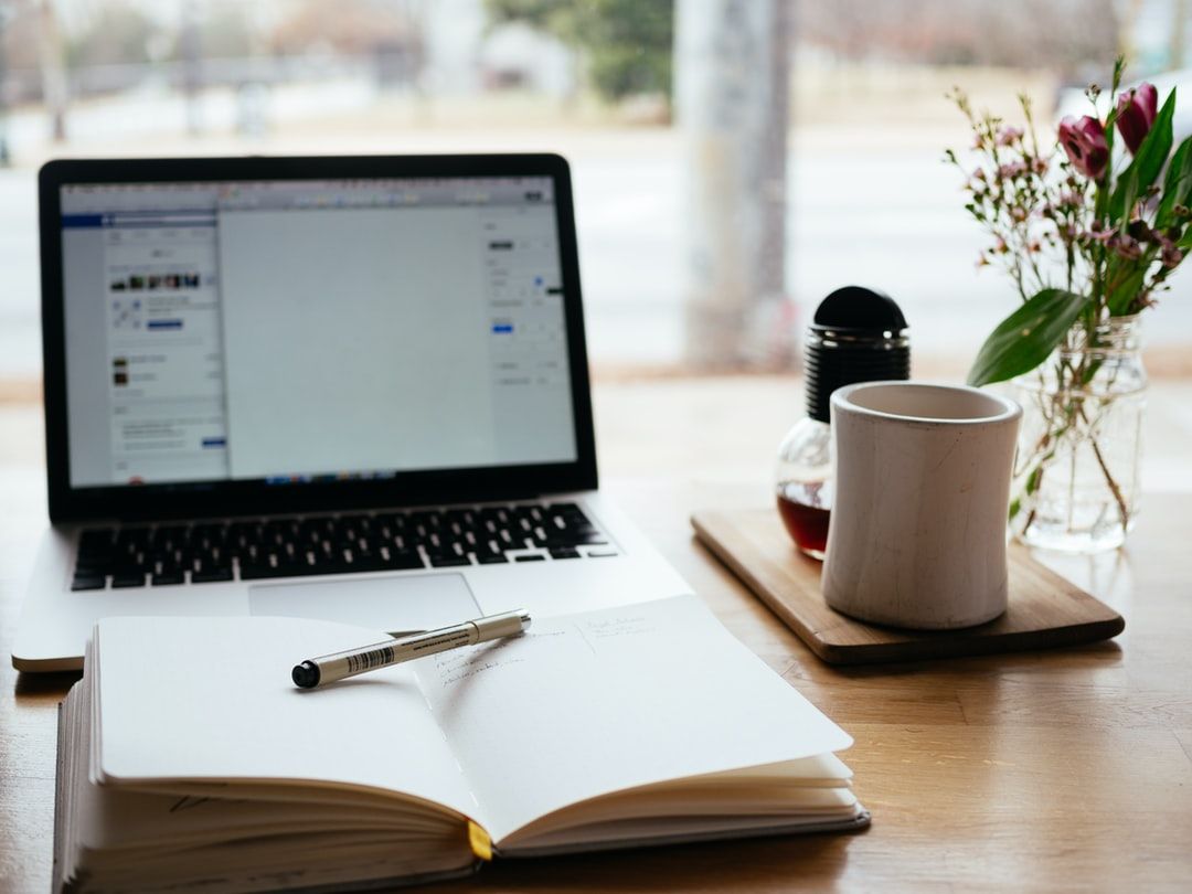 desk with an open journal and pen sitting on top if, above it on desk an open laptop, on right side of laptop and book a cutting board with cup and a tea diffuser